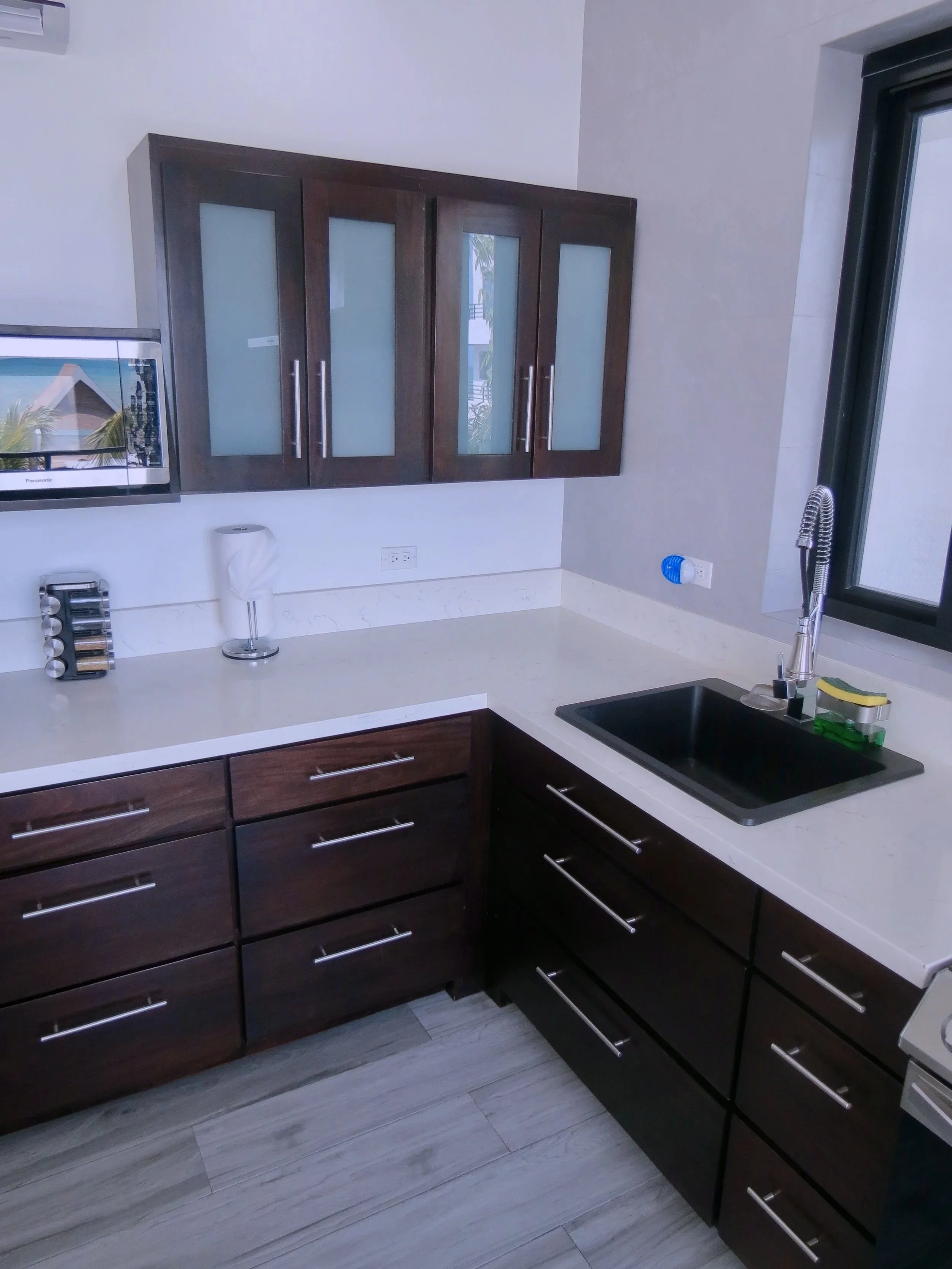 Modern kitchen with dark wood cabinets, a white countertop, a black sink, and a window with a view of outdoor trees and buildings.  Unit S2, Caye Caulker,  Blu Zen Belize
