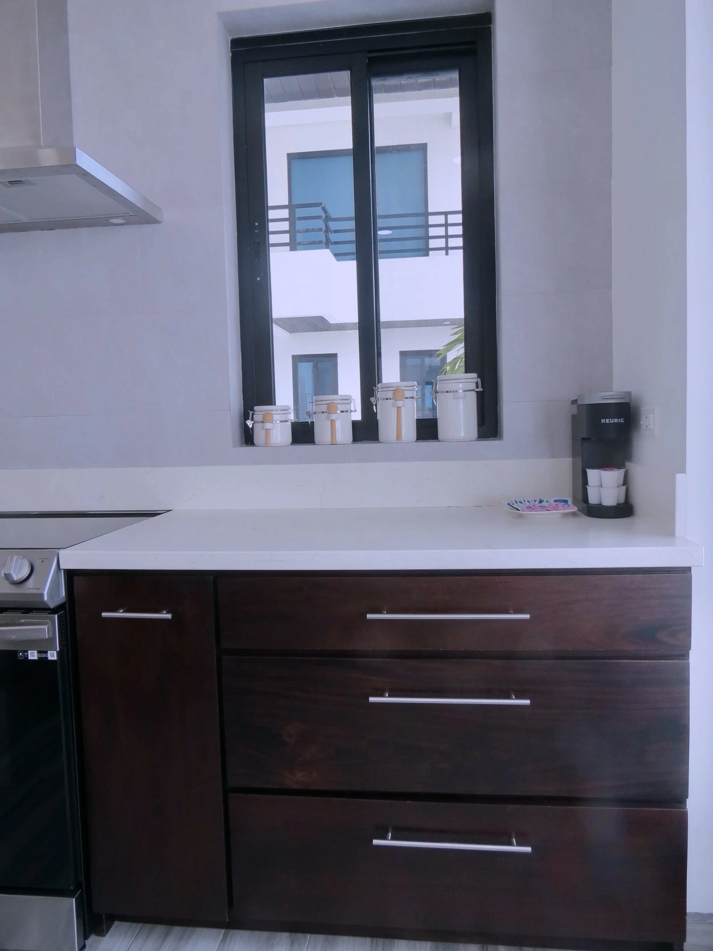 Kitchen with dark wood cabinets, a white countertop, and a window showing an exterior balcony with blue doors. Items on the countertop include white canisters and a Keurig coffee maker.