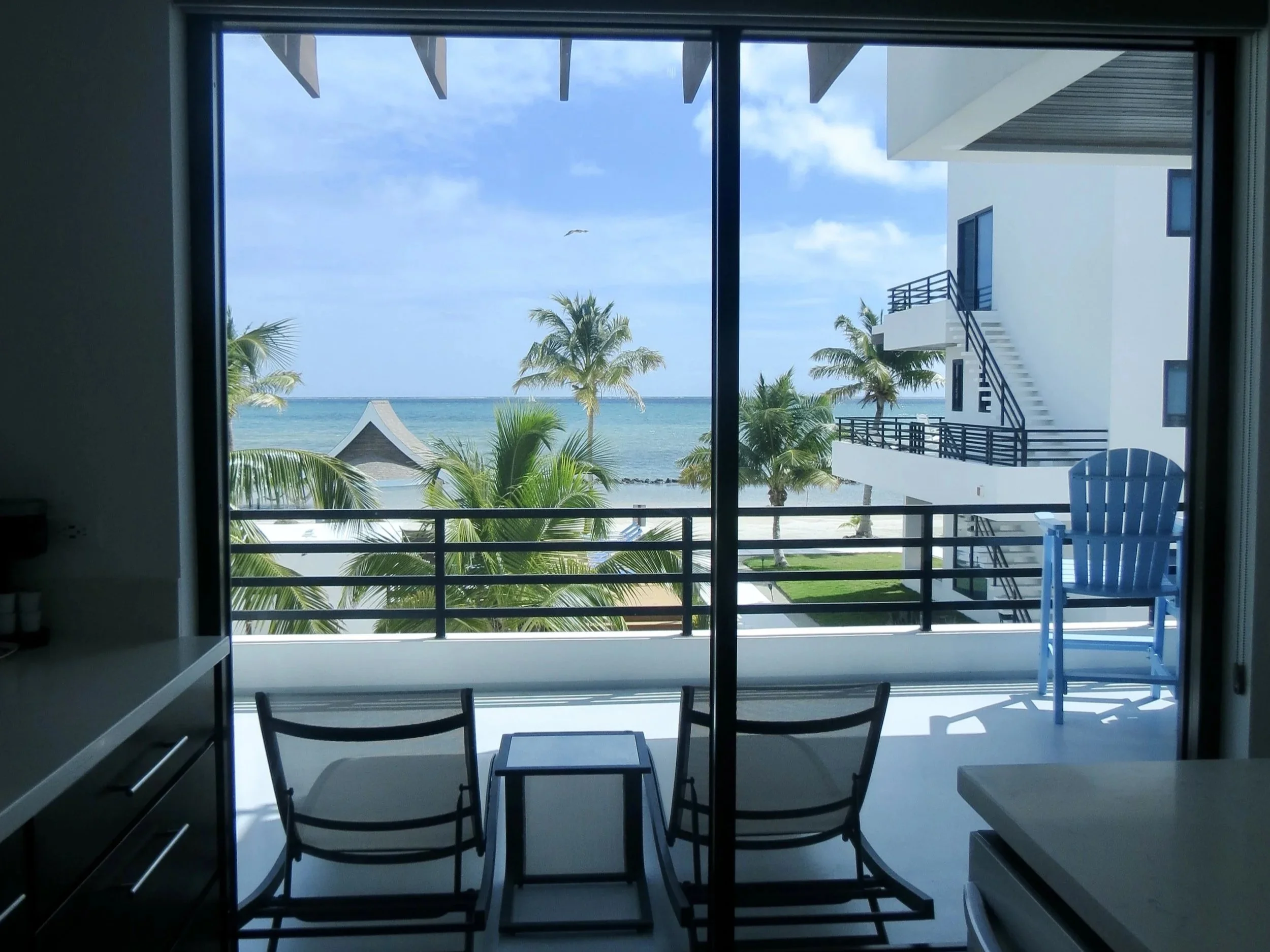 View of a tropical beach through a sliding glass door, with palm trees, a thatched roof hut, and ocean in the background; balcony with blue chair outside and chairs inside.