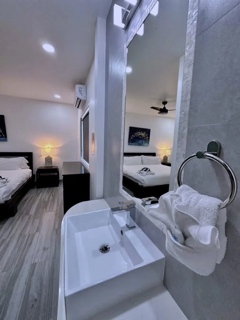 Primary bedroom viewed from the ensuite bathroom, showing a king-size bed, a bedside table with a lamp, and a framed painting on the wall.  Bathroom sink with a mirror and a towel ring with a folded towel.  Unit S2, Caye Caulker,  Blu Zen Belize