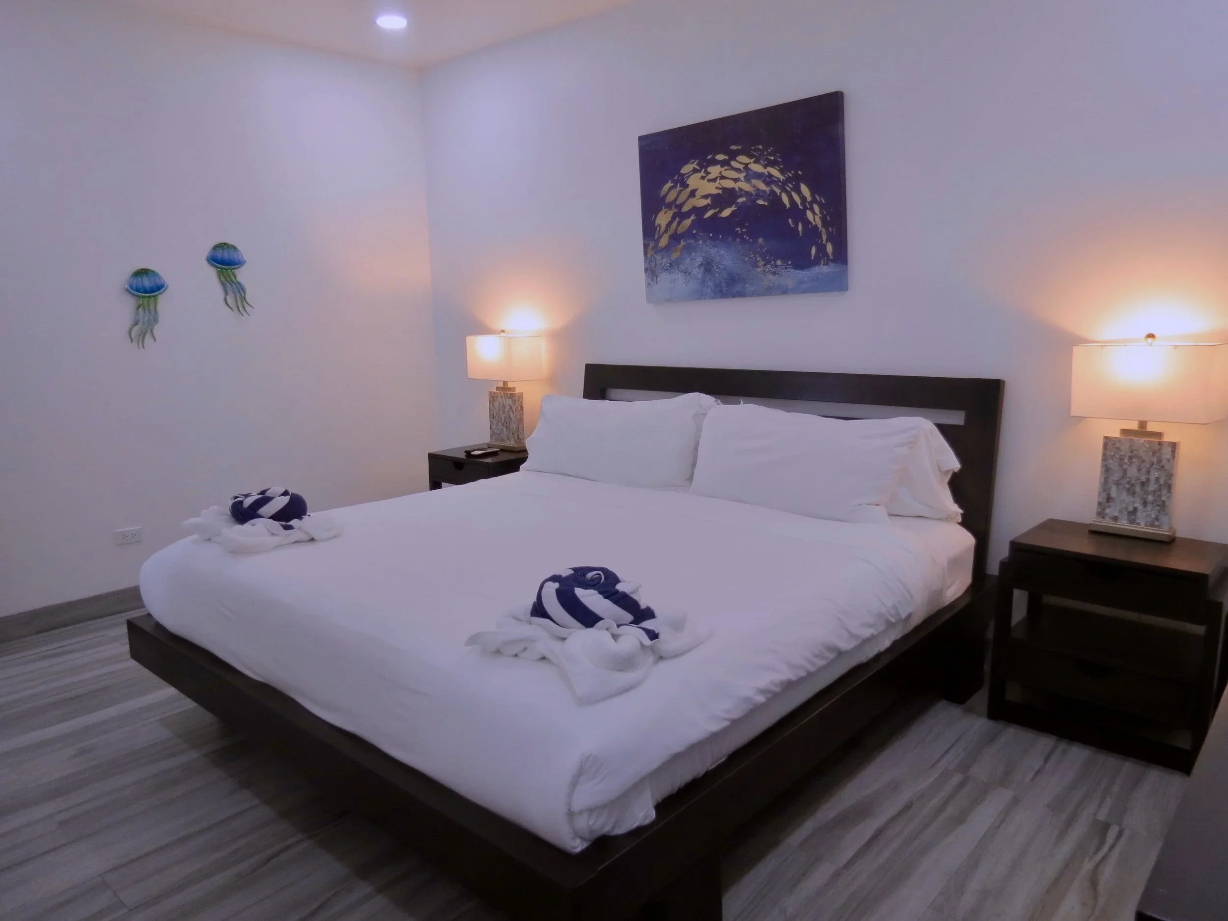 Primary bedroom with a large king-size bed, two nightstands with lamps, ocean-themed artwork, jellyfish wall decorations, and towels folded into swan shapes.  Unit S2, Caye Caulker,  Blu Zen Belize