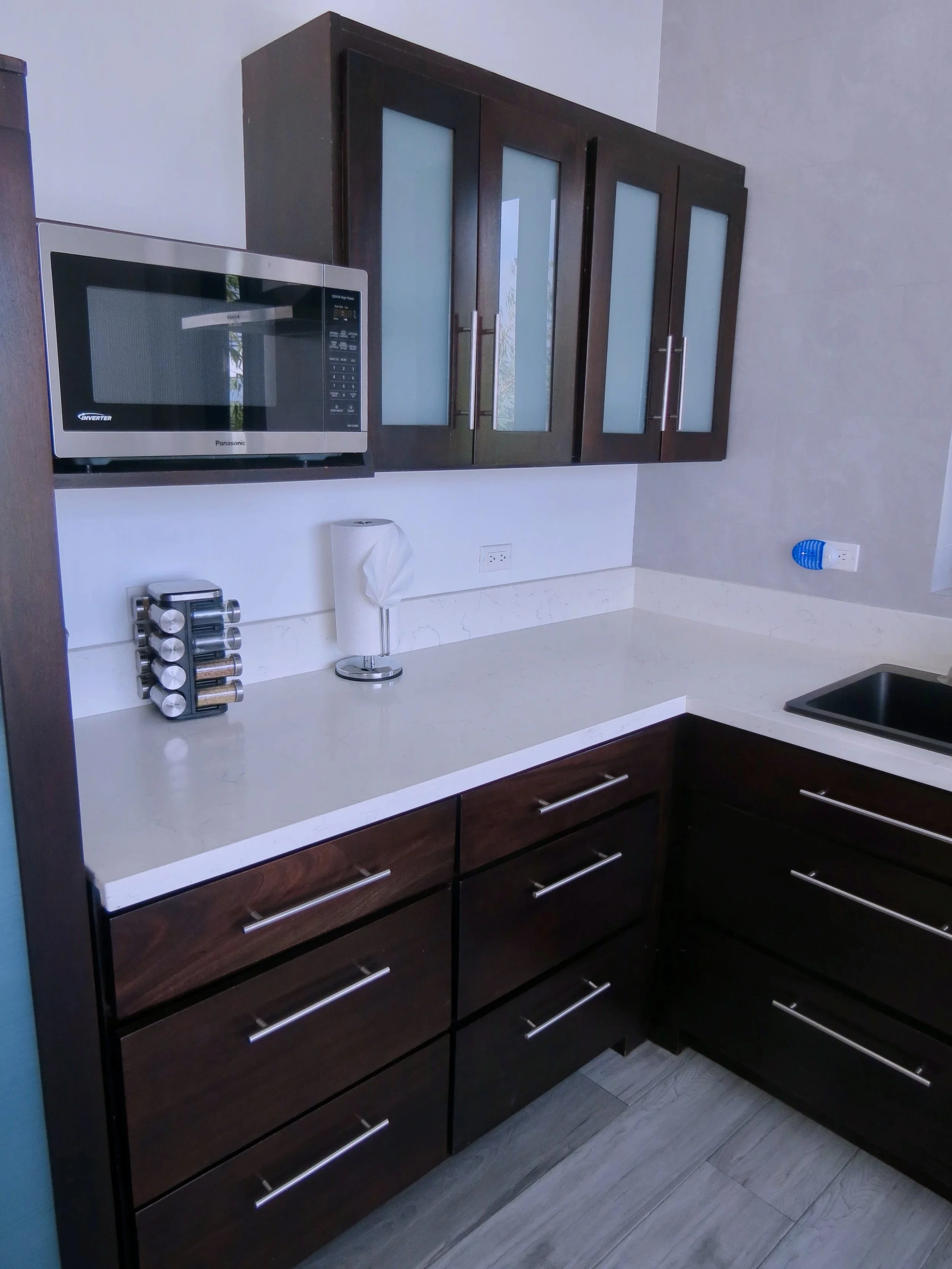 Kitchen with dark wood cabinets, white countertop, microwave, spice rack, paper towel holder, electrical outlet, and black sink.  Unit S2, Caye Caulker,  Blu Zen Belize