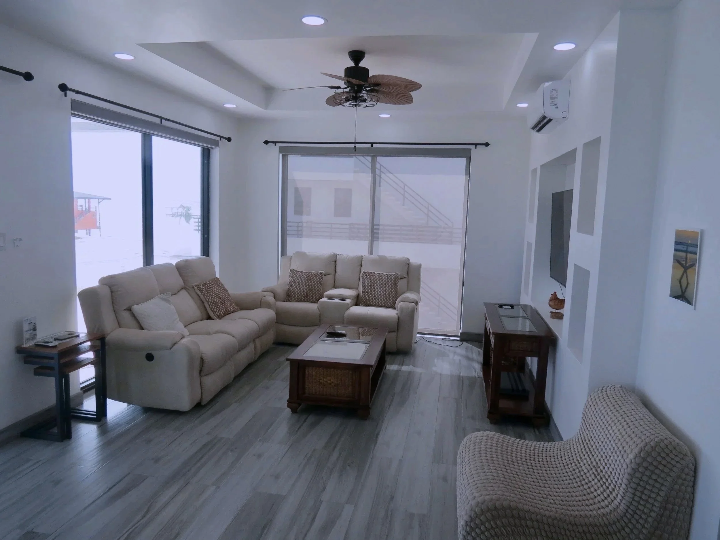 Living room with white walls, large windows with blinds, beige furniture including a sofa and an armchair, wooden coffee table, side tables, ceiling fan, and wall-mounted TV.  Unit S2, Caye Caulker,  Blu Zen Belize