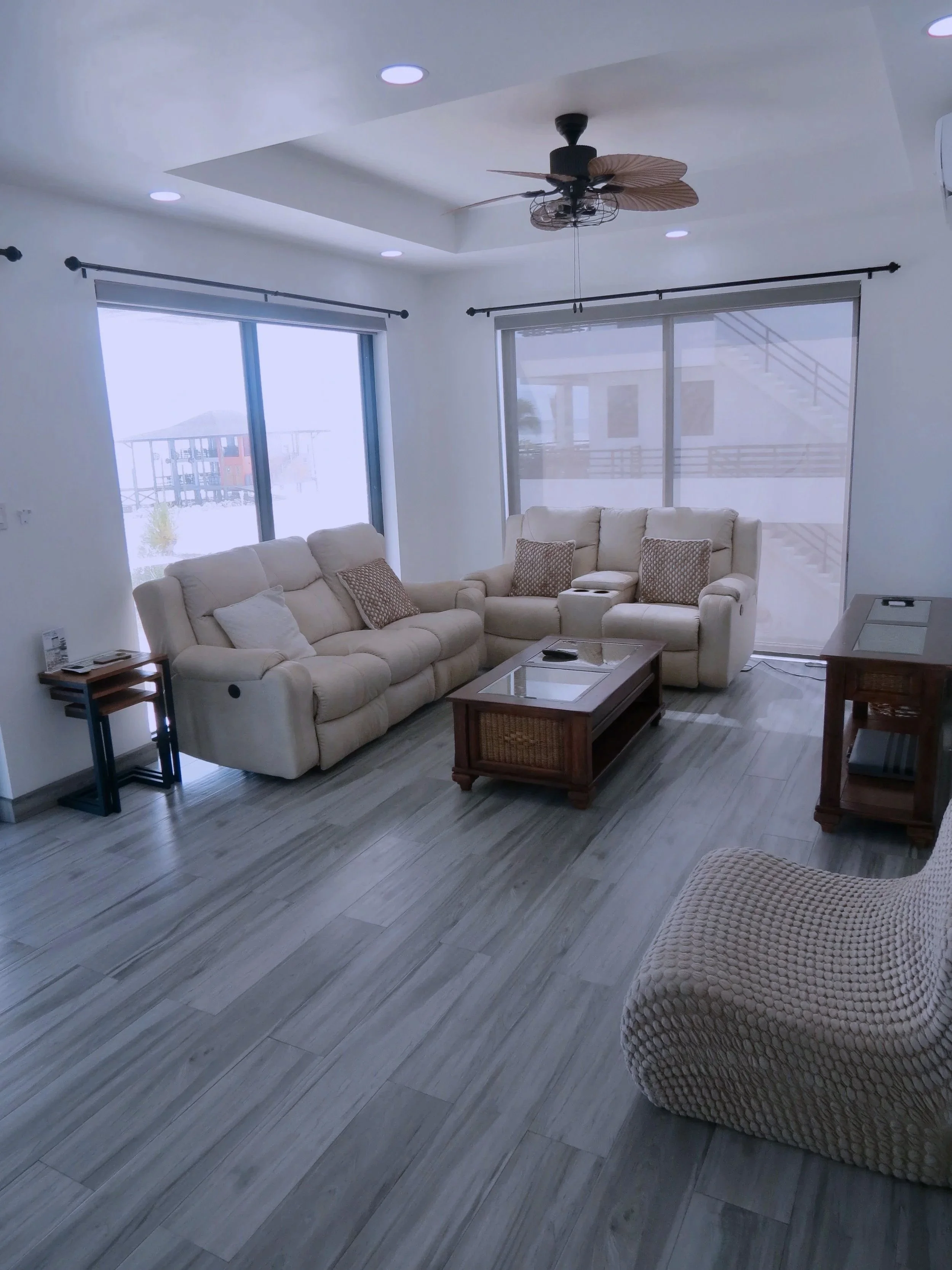 Bright living room with white sofas, a glass-top coffee table, a wicker chair, large windows, and a ceiling fan, with ocean view outside.  Unit S2, Caye Caulker,  Blu Zen Belize
