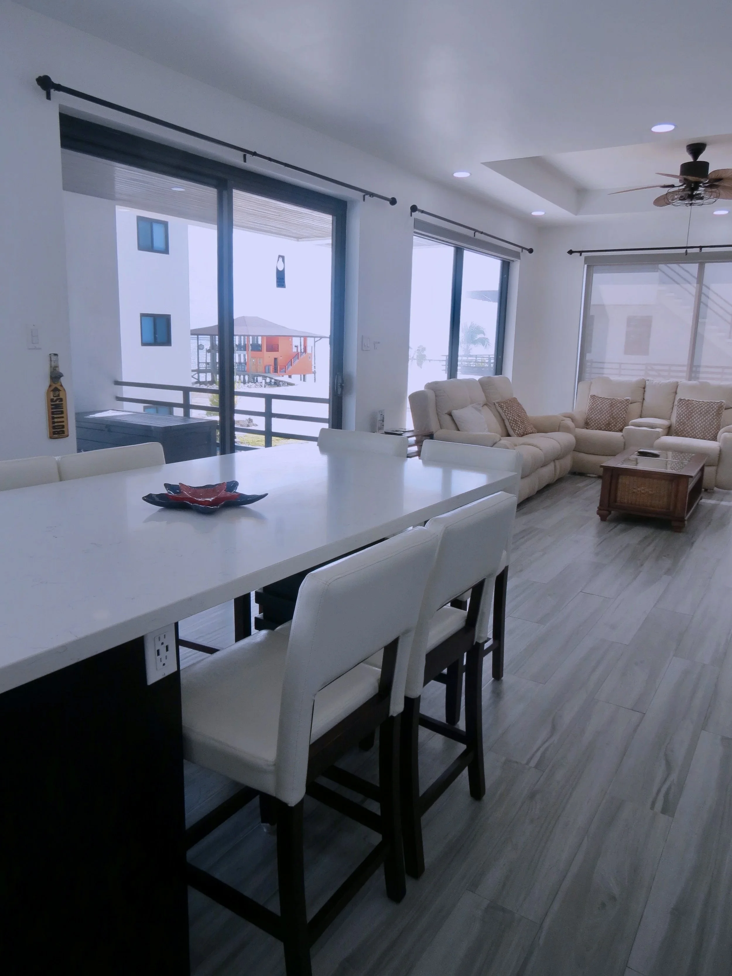 Bright living room with large sliding glass doors, beige sectional sofa with pillows, coffee table, ceiling fan, and a dining area with white chairs and a white table.  Unit S2, Caye Caulker,  Blu Zen Belize