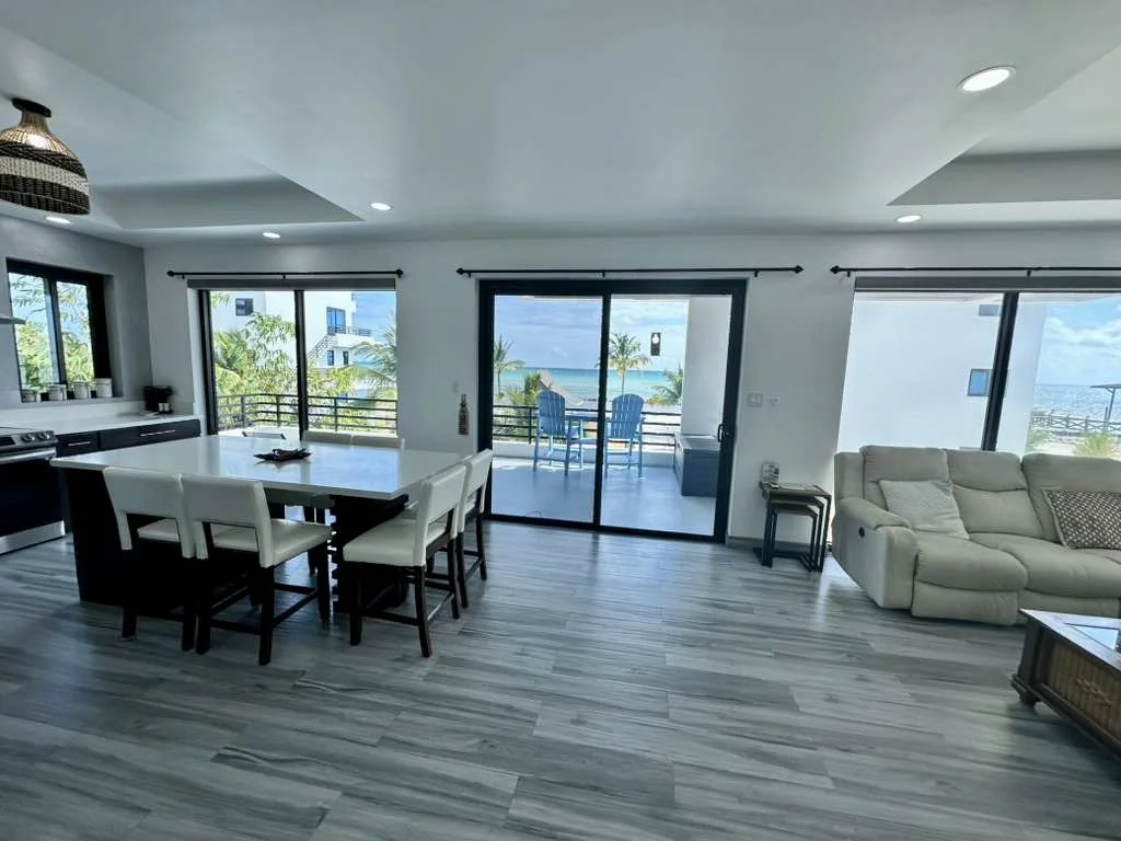 Open-concept living and dining area with large windows and sliding glass doors revealing a balcony with outdoor seating and a view of the ocean and palm trees.  Unit S2, Caye Caulker,  Blu Zen Belize