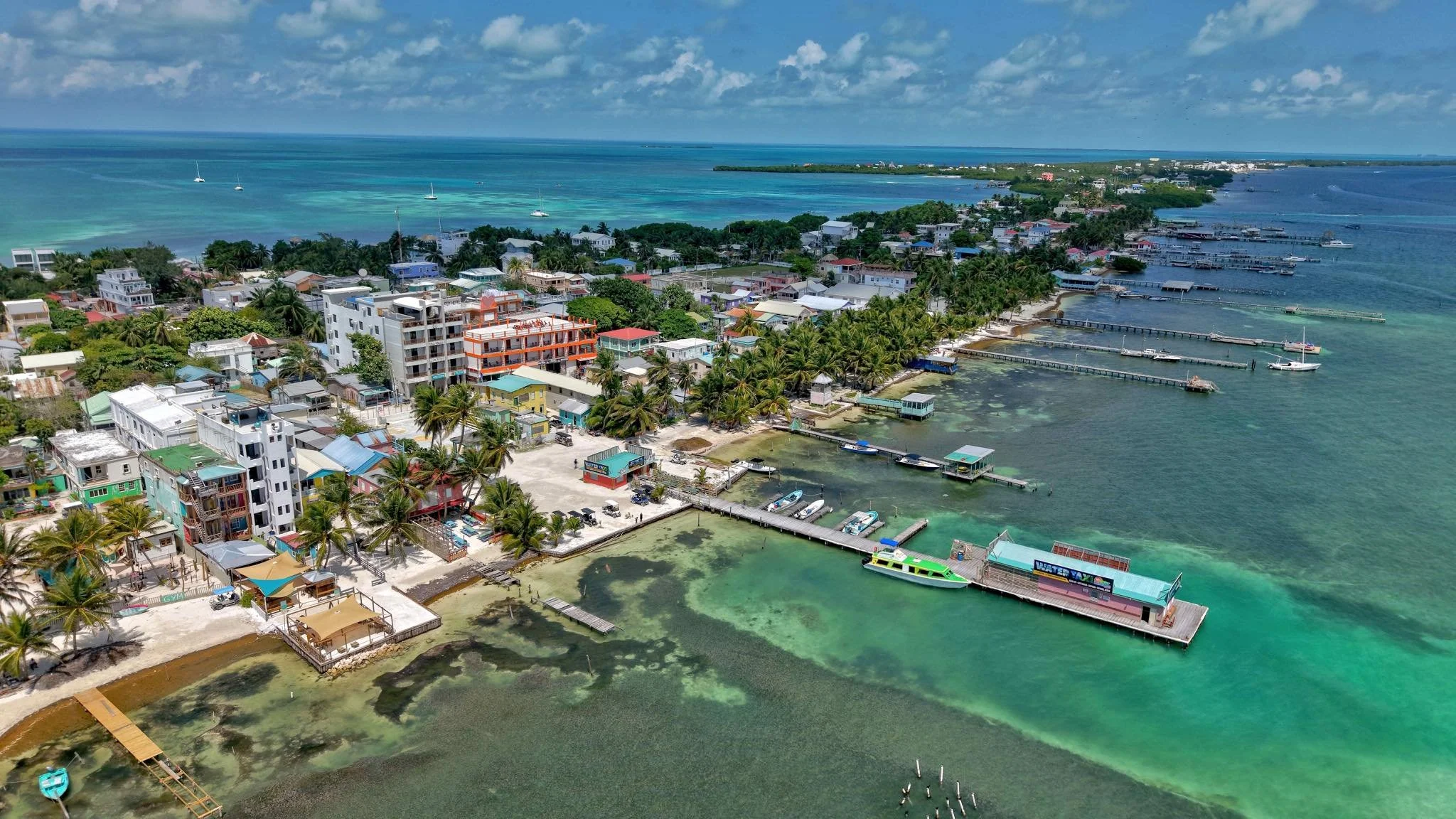 Aerial view of a tropical seaside town with colorful buildings, palm trees, and boats at a marina, with turquoise waters and a distant coastline under a partly cloudy sky.  Unit S2 Blu Zen Belize