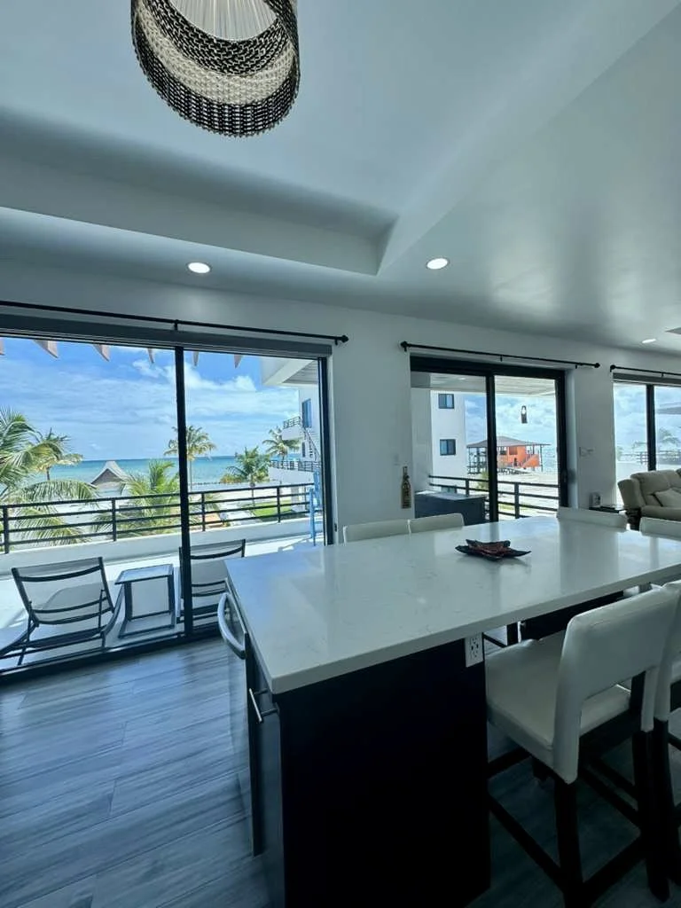 Indoor view of a modern living and dining area with large sliding glass doors showing a beach view with palm trees and ocean.  Unit S2, Caye Caulker,  Blu Zen Belize