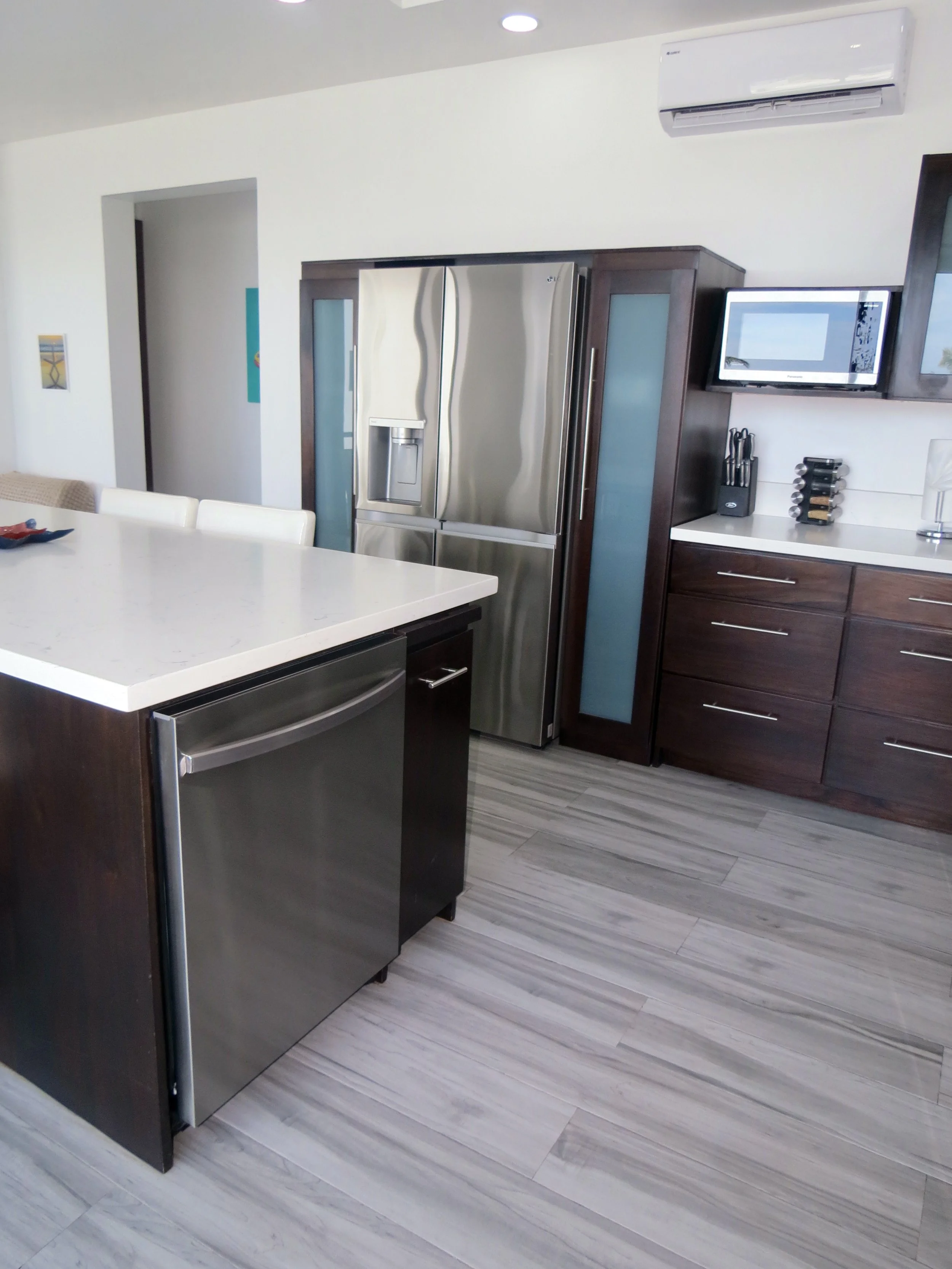 Modern kitchen with stainless steel appliances, including dishwasher, dark wooden cabinets, white countertop, and light-colored tile flooring.  Unit S2, Caye Caulker,  Blu Zen Belize