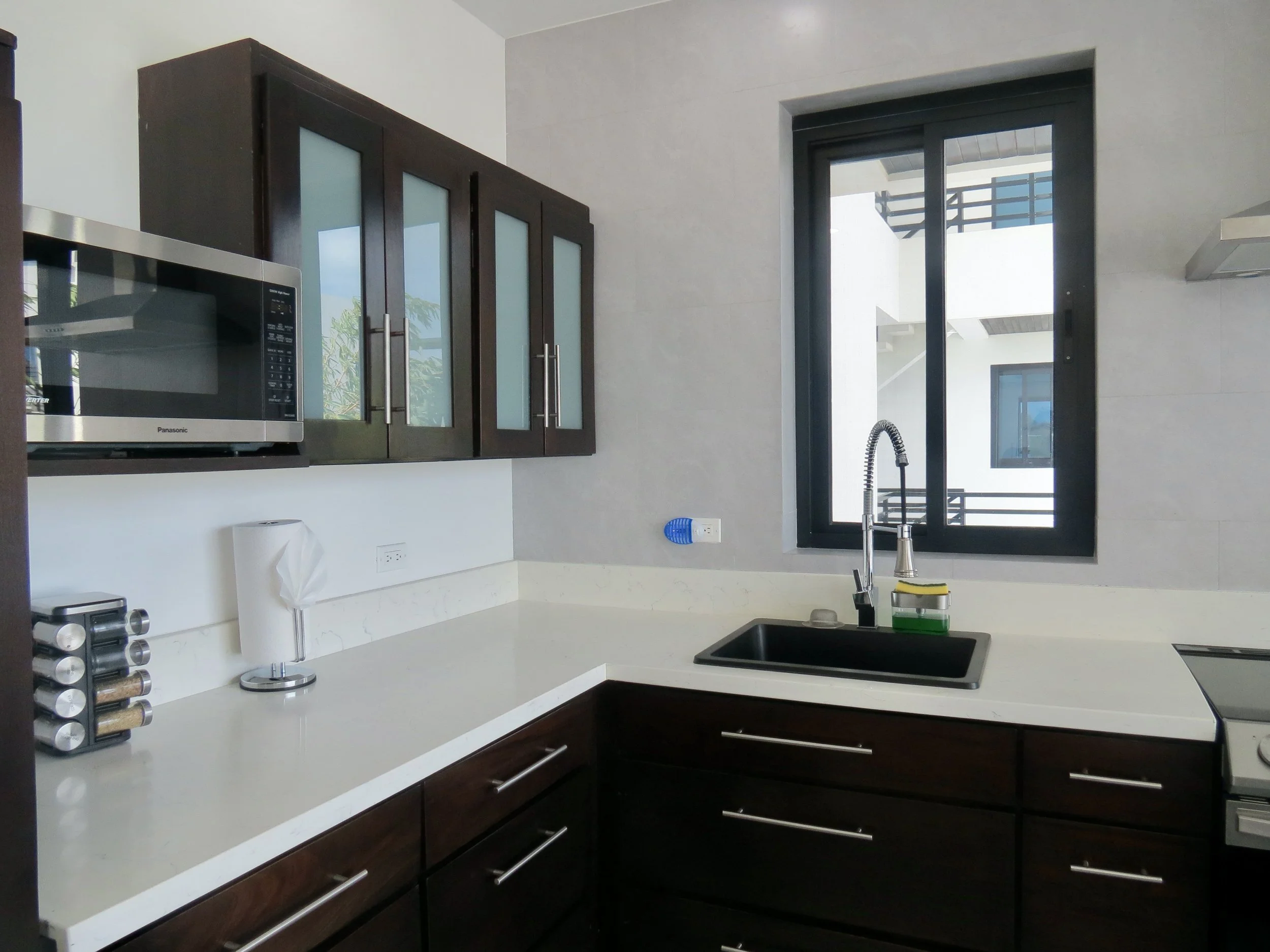 Modern kitchen with dark wood cabinets, white countertops, a black sink, a window with black frame, and a microwave on the left.  Unit S2, Caye Caulker,  Blu Zen Belize
