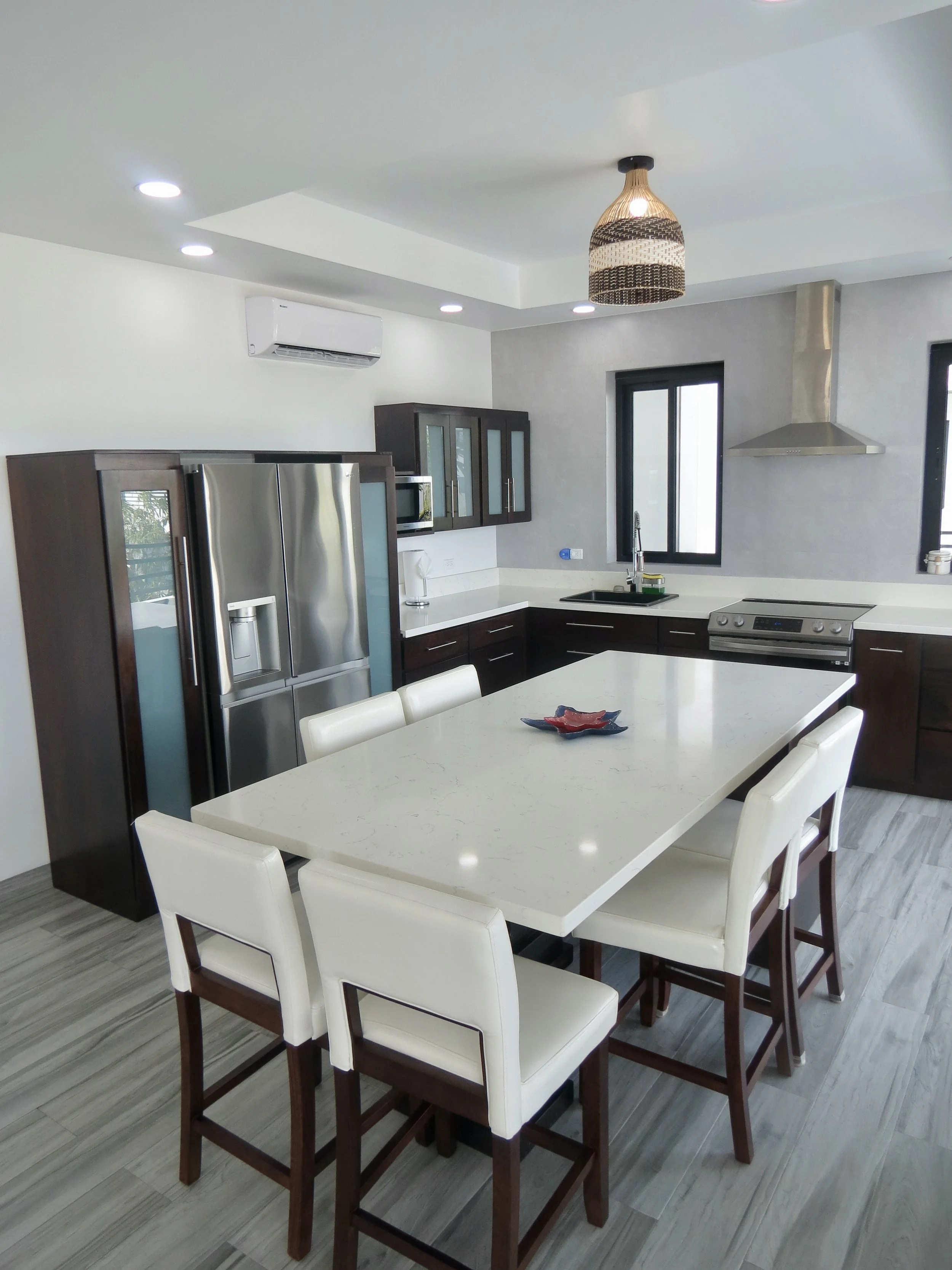 Modern kitchen with a white island table, six white chairs, dark wood cabinetry, stainless steel refrigerator, stove, microwave, and range hood, gray flooring, and two windows with black frames.  Unit S2, Caye Caulker,  Blu Zen Belize
