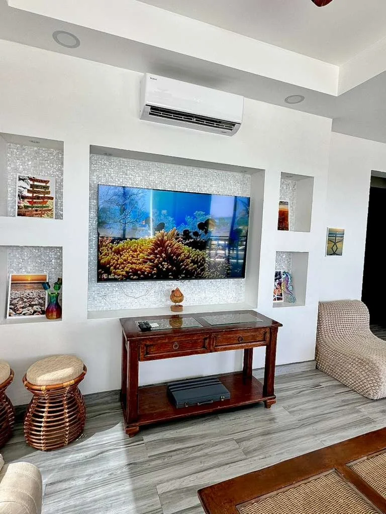 Living room with wall-mounted TV displaying an underwater coral scene, wooden console table below with remote controls, woven stools to the left, and artwork on shelves set in a white wall. Air conditioner above the TV and wood-textured-loo flooring.