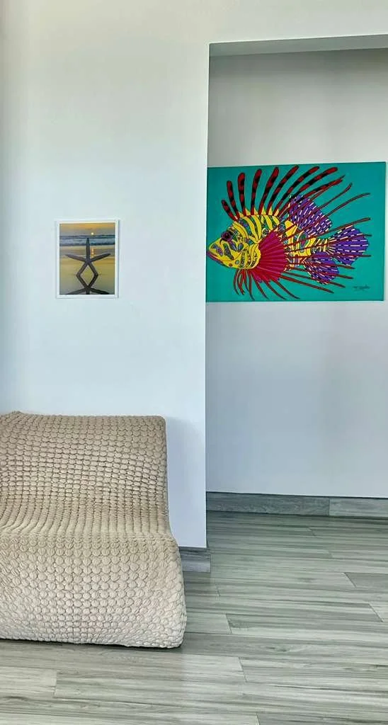 Interior of a room with light-colored walls, a textured beige sofa, and two colorful pieces of artwork: one small landscape and one large, vibrant fish painting.