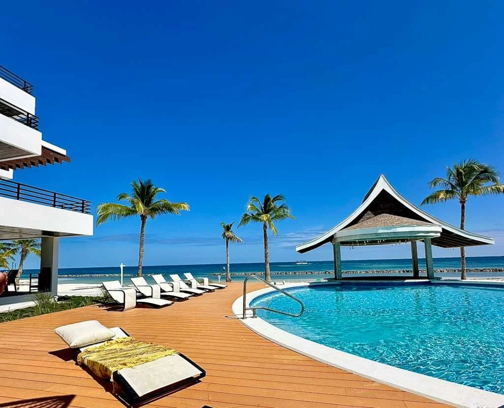 A tropical pool area with poolside chairs, a wooden deck, palm trees, a pavilion, and an ocean view with a clear blue sky.  Phase 3 Swimming Pool in front of Unit S2 Blu Zen Belize