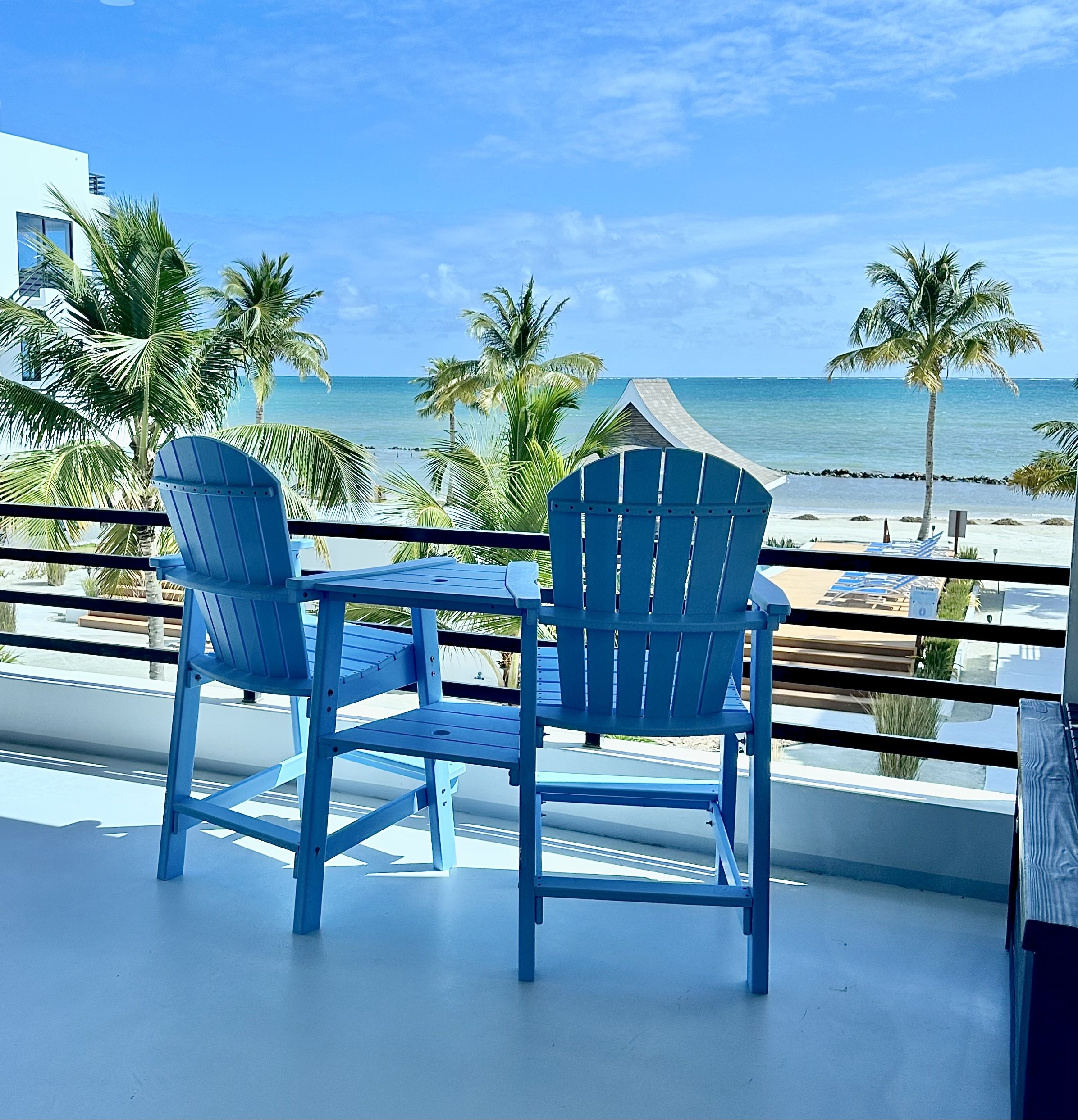 Two blue Adirondack chairs on a balcony overlooking a tropical beach with palm trees, a thatched hut, and the ocean under a partly cloudy sky.  Private Balcony, Unit S2 Blu Zen Belize