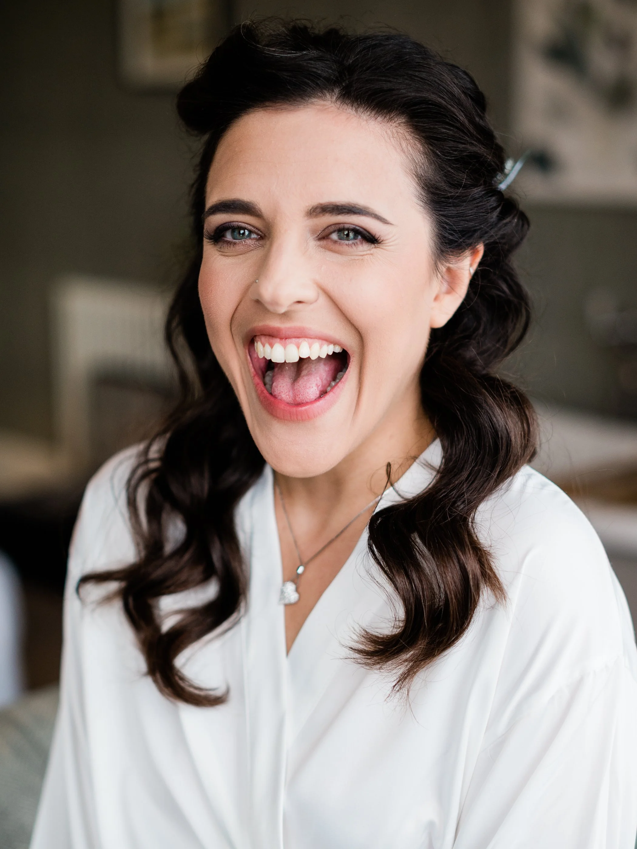 A woman with dark wavy hair, wearing a white top and a silver heart-shaped pendant necklace, smiling widely with her mouth open.