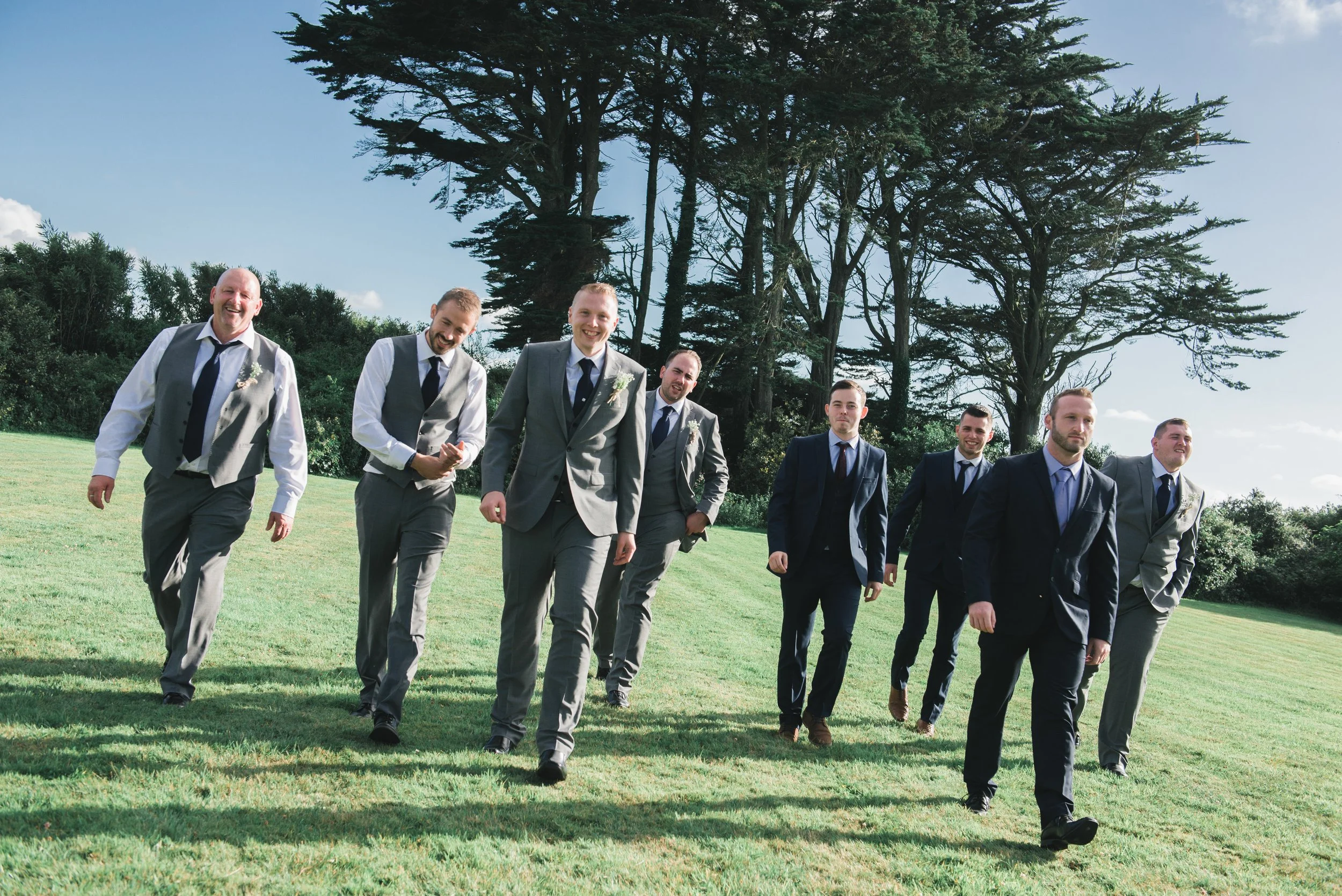 A group of eight men dressed in suits and vests walking across a grassy field with large trees in the background, smiling and enjoying themselves.