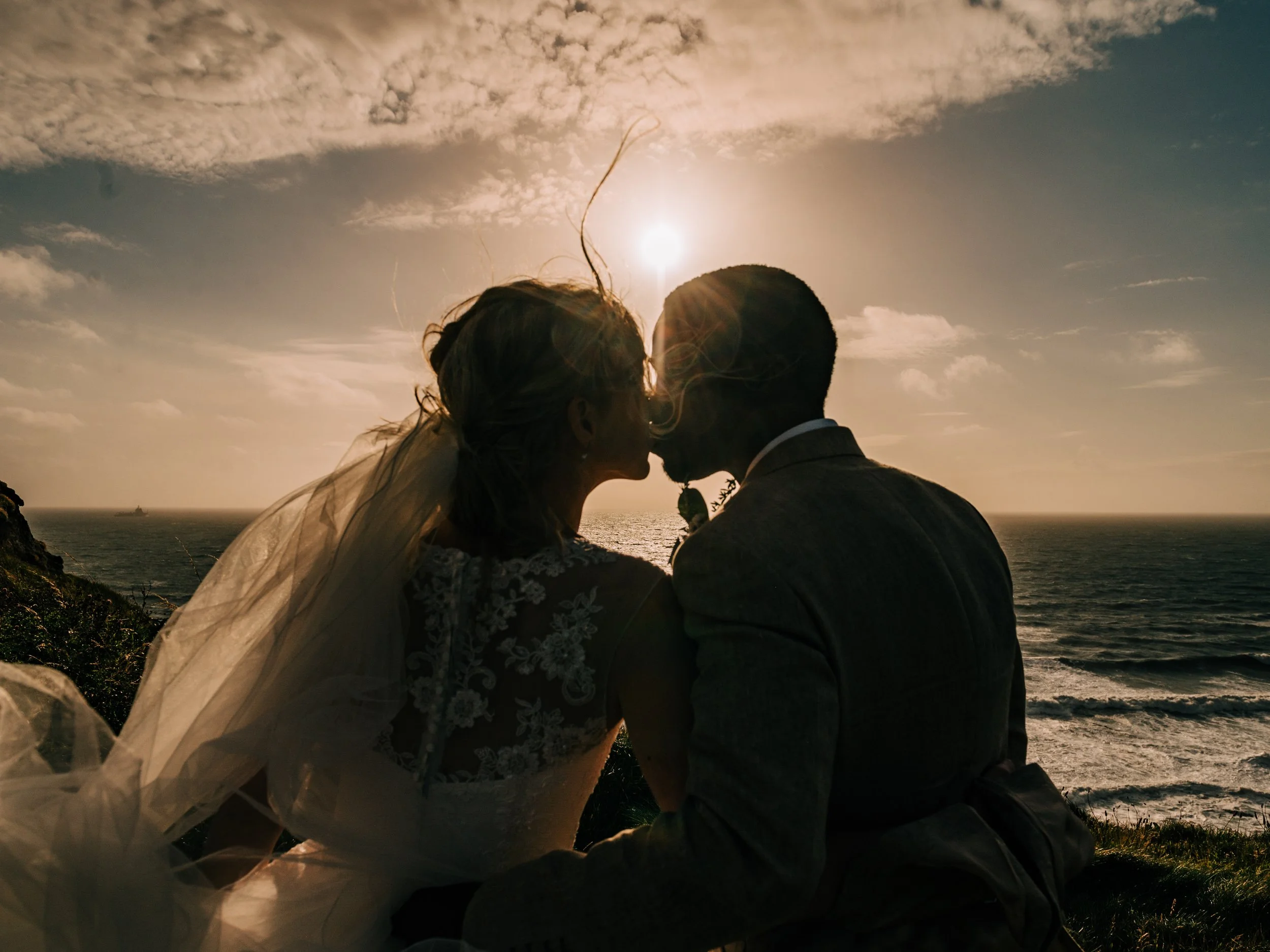A couple in wedding attire sharing a kiss at sunset by the ocean, with the sun setting behind them and waves in the background.