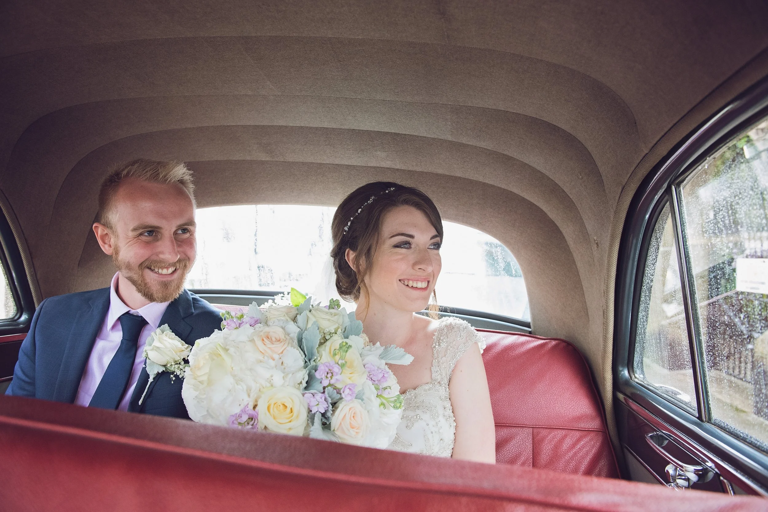 Bride and groom sitting in the back of a vintage car, smiling, with the bride holding a bouquet of white, pink, and purple flowers, during their wedding celebration.