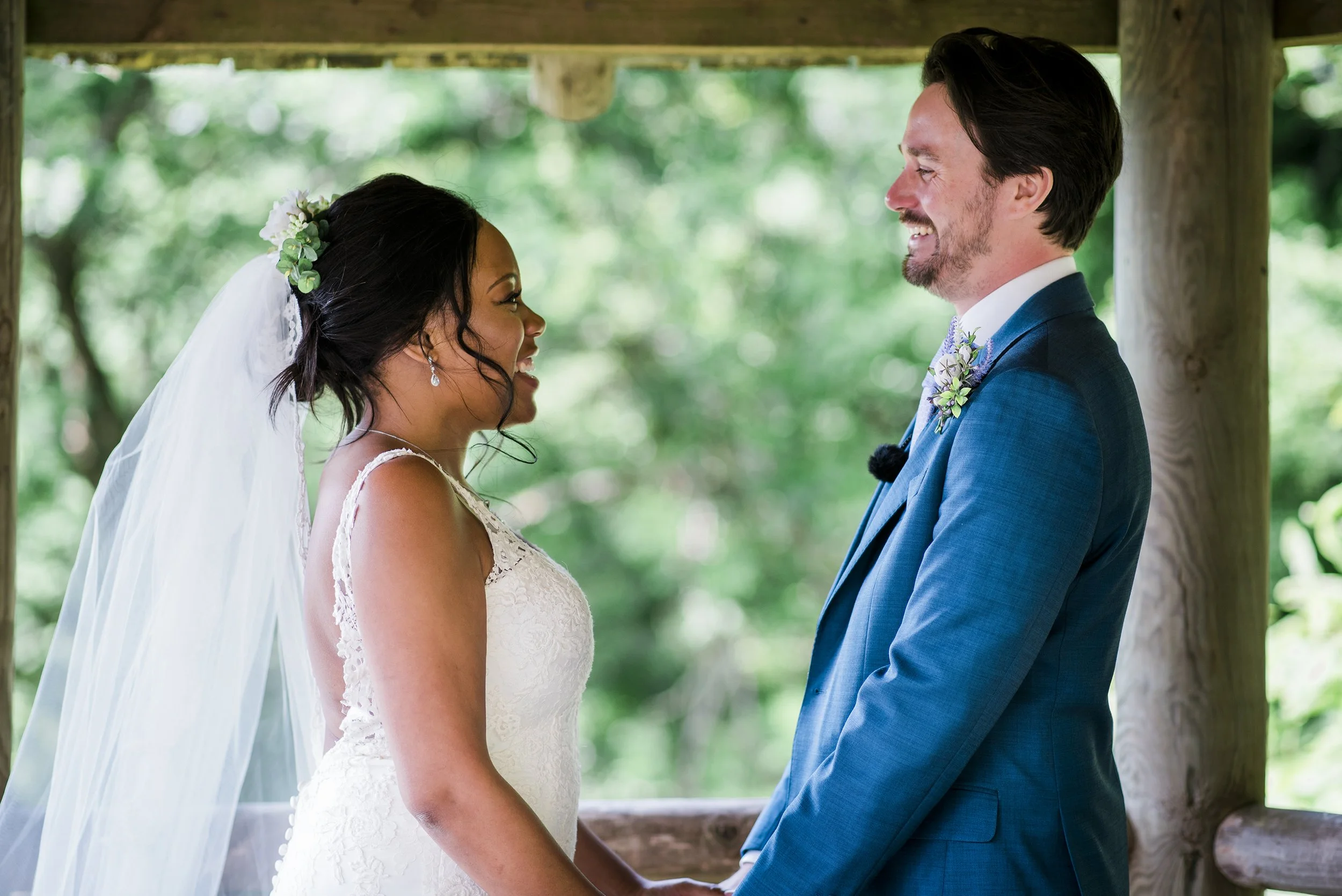 A bride and groom holding hands and smiling at each other during their wedding ceremony outdoors with green trees in the background.