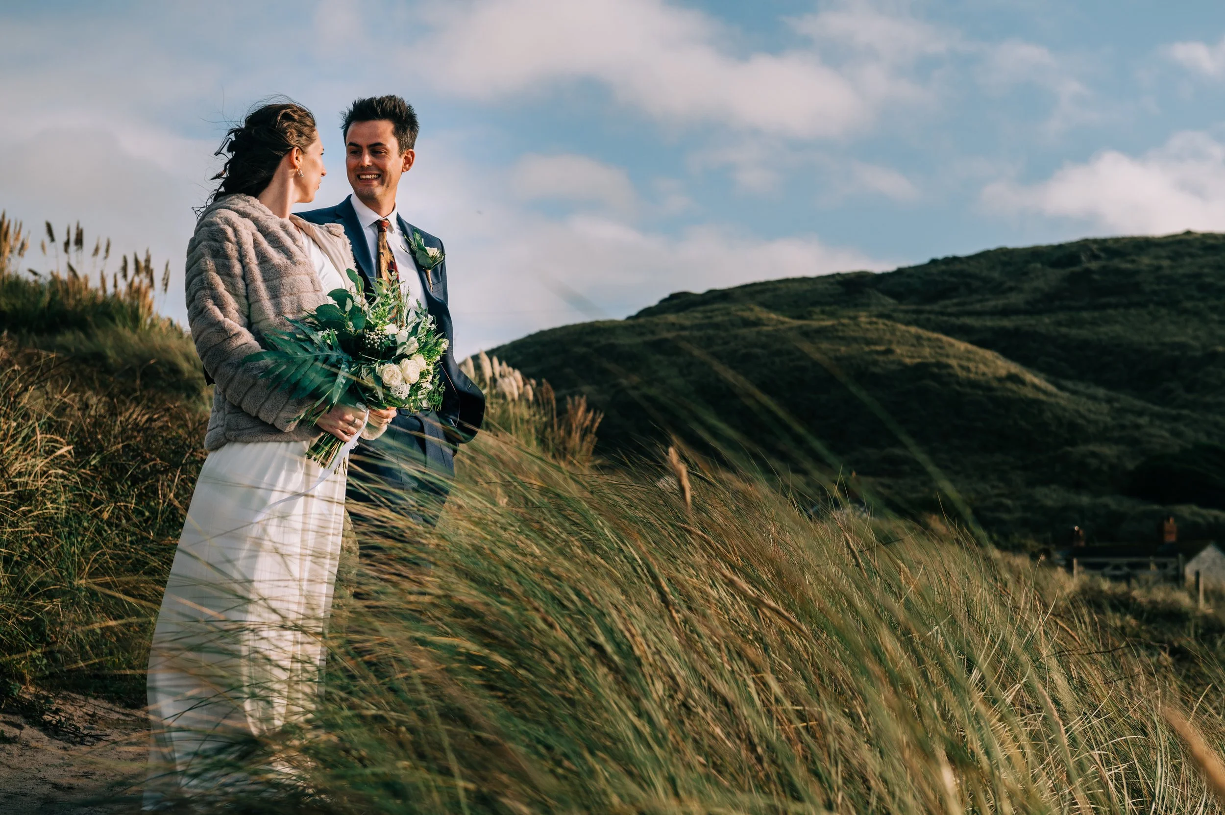 A couple in wedding attire standing outdoors amidst tall grass with hills in the background, holding a bouquet of flowers, and smiling at each other.