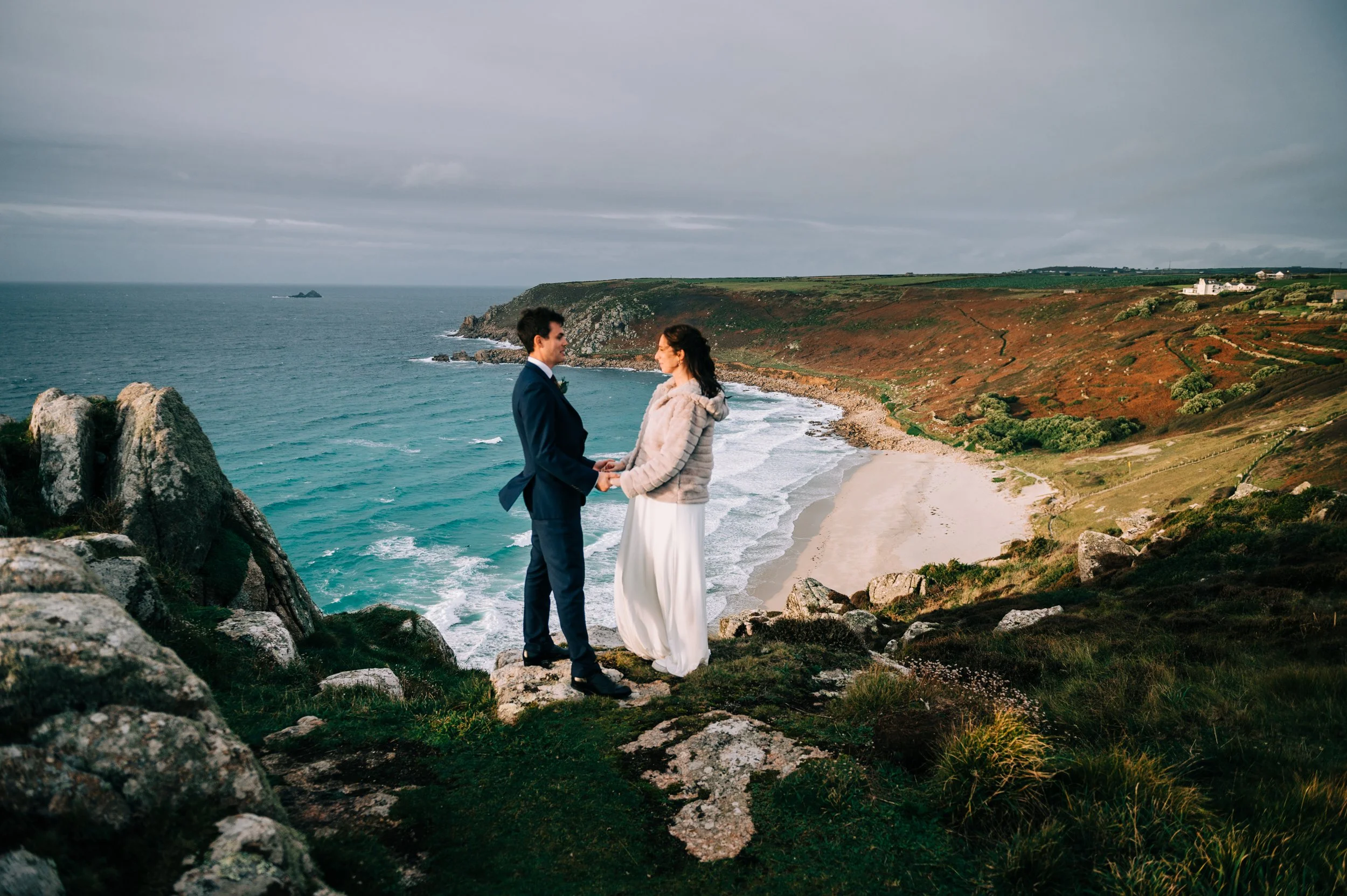 A couple getting married on a cliff overlooking a beach and ocean, holding hands and facing each other.