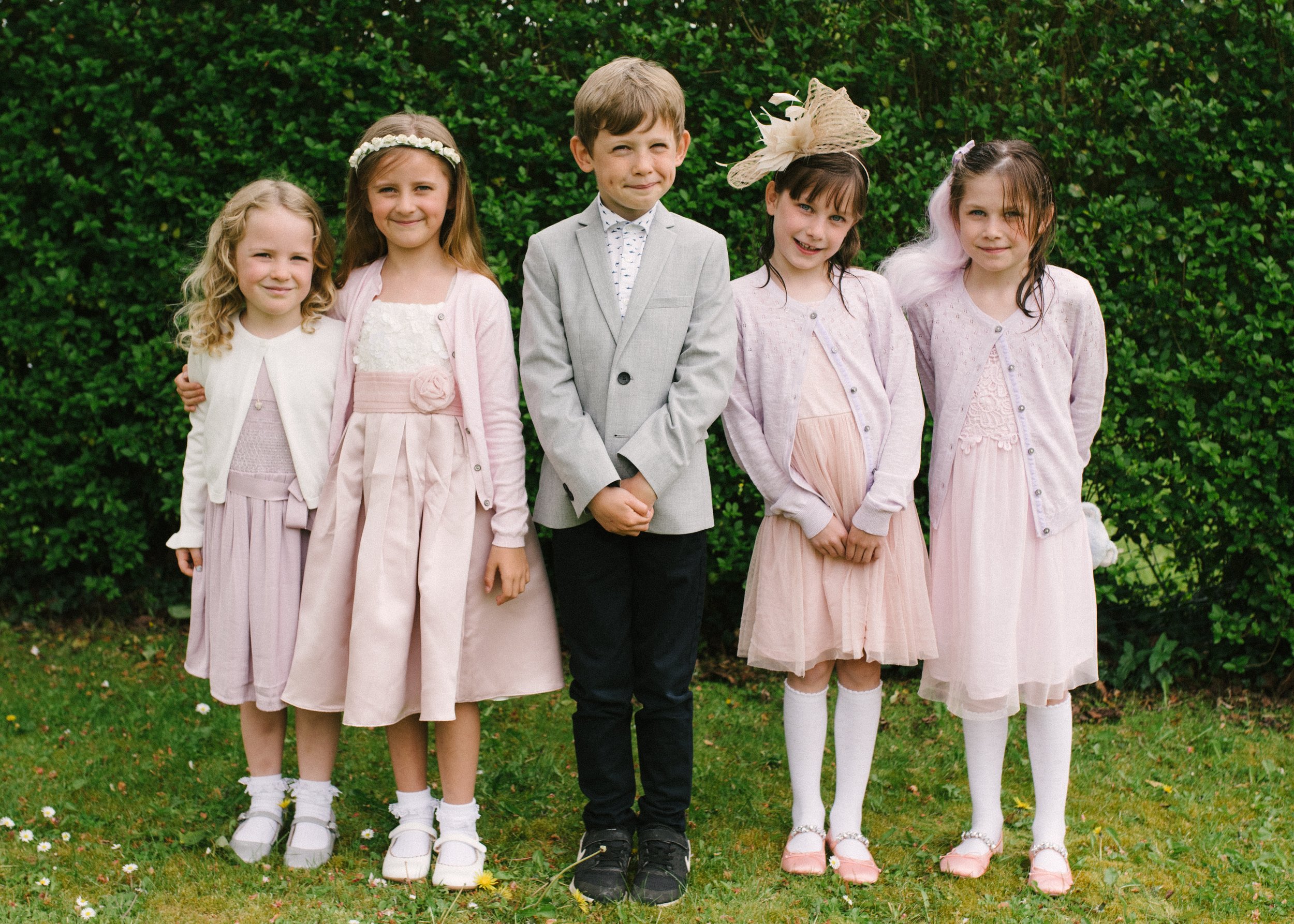 Group of five young children, four girls and one boy, dressed in pastel and neutral colored clothing, standing outdoors on grass in front of green bushes.