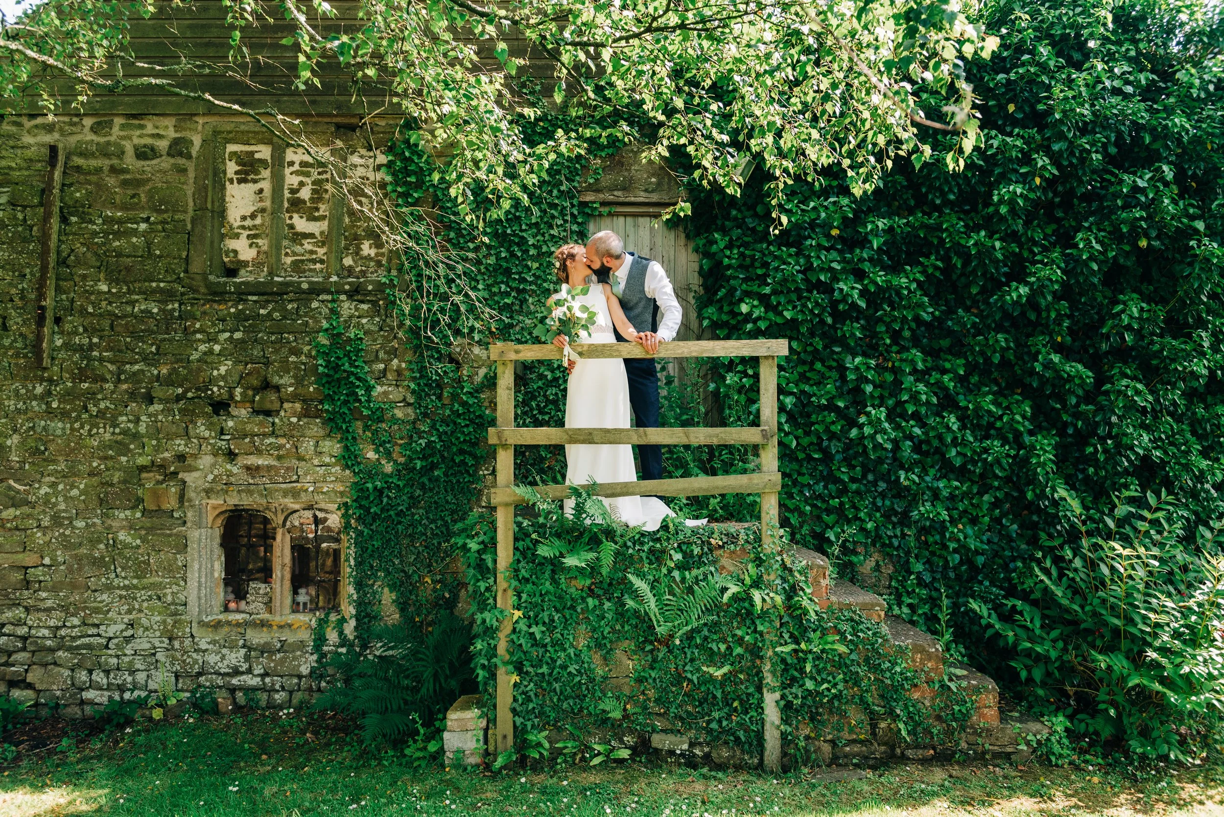 A bride and groom sharing a kiss on a small wooden balcony surrounded by green ivy and foliage, with an old brick building in the background.