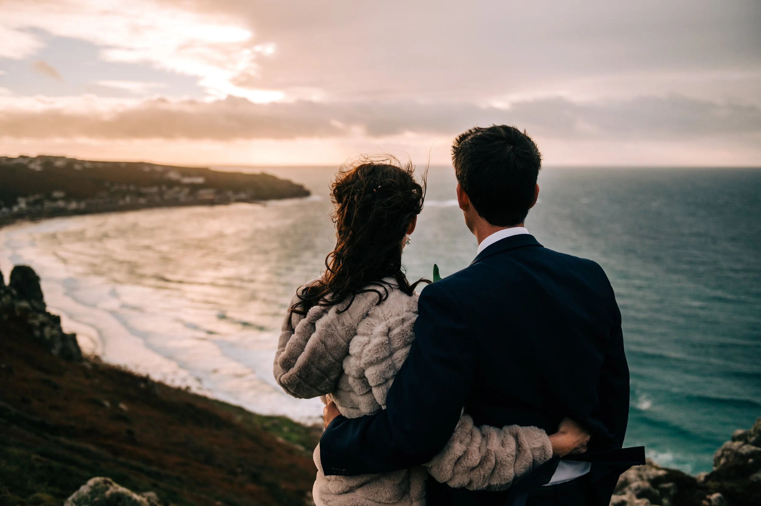 A couple in formal attire embracing and looking at the ocean during sunset.