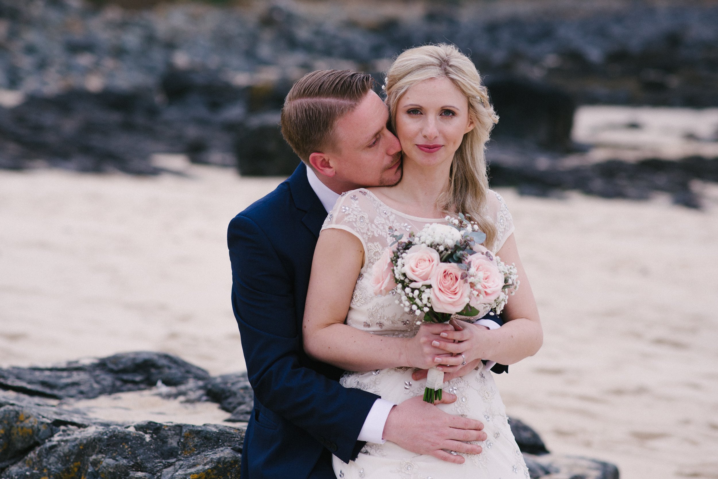 A couple dressed in wedding attire, with the groom kissing the bride on her cheek, standing on rocks near the water on a beach.