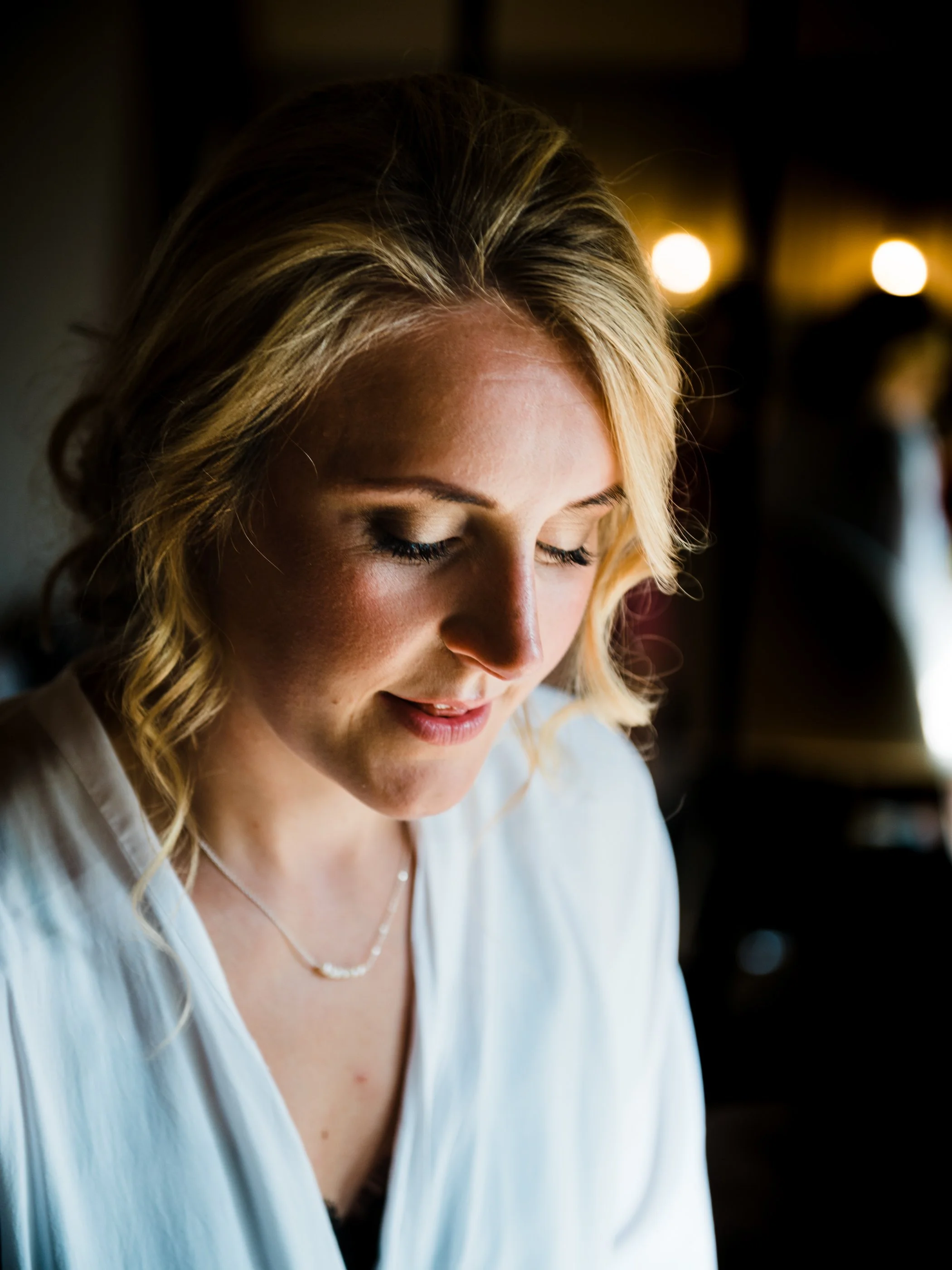 Close-up of a young woman with blonde wavy hair, looking down and smiling gently, wearing a white top and a delicate necklace, indoors with warm lighting.
