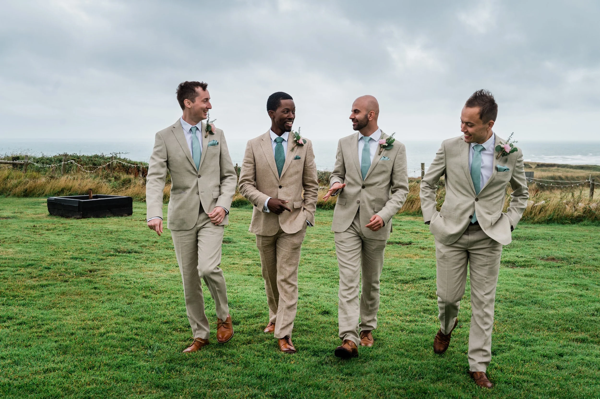 Four men in beige suits walking on a grassy outdoor area, smiling and talking, with a cloudy sky and ocean in the background.