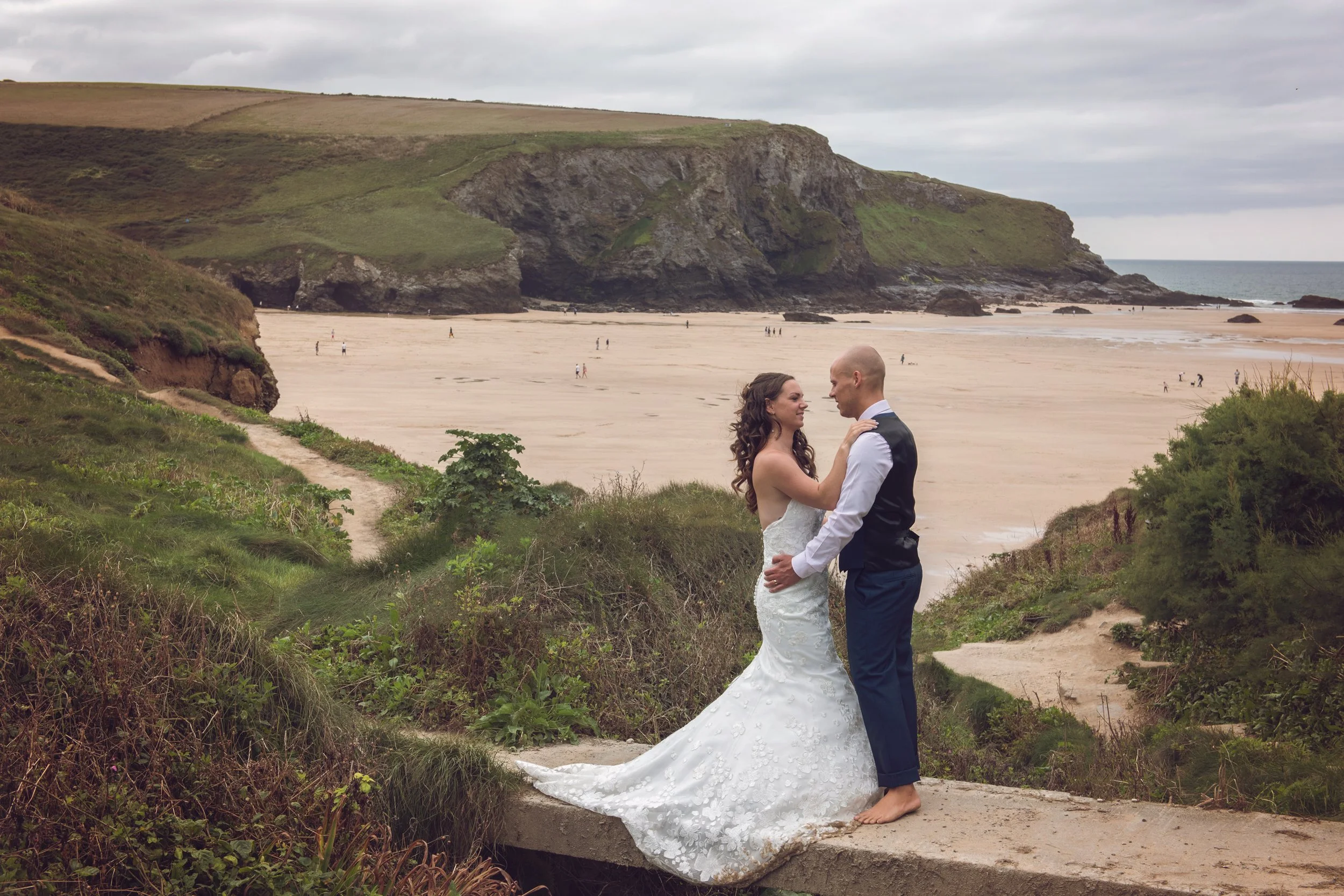 A bride and groom standing close together on a grassy path by the beach, embracing and gazing into each other's eyes with a large sandy beach, cliffs, and the ocean in the background.