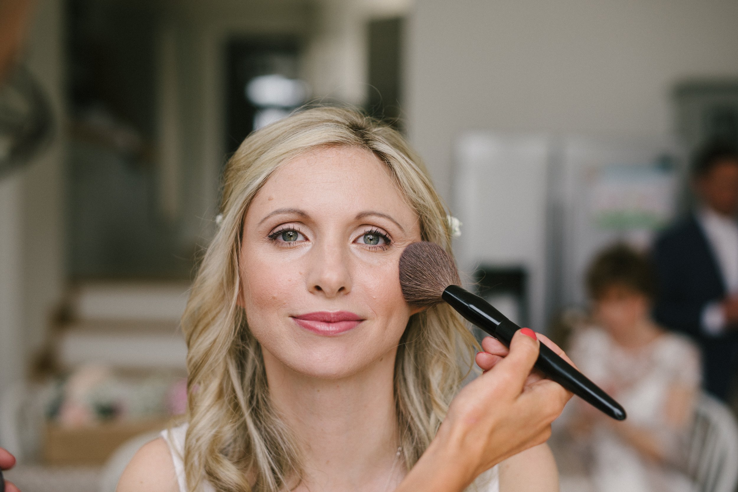 A woman with blonde hair gets her makeup done using a large powder brush.