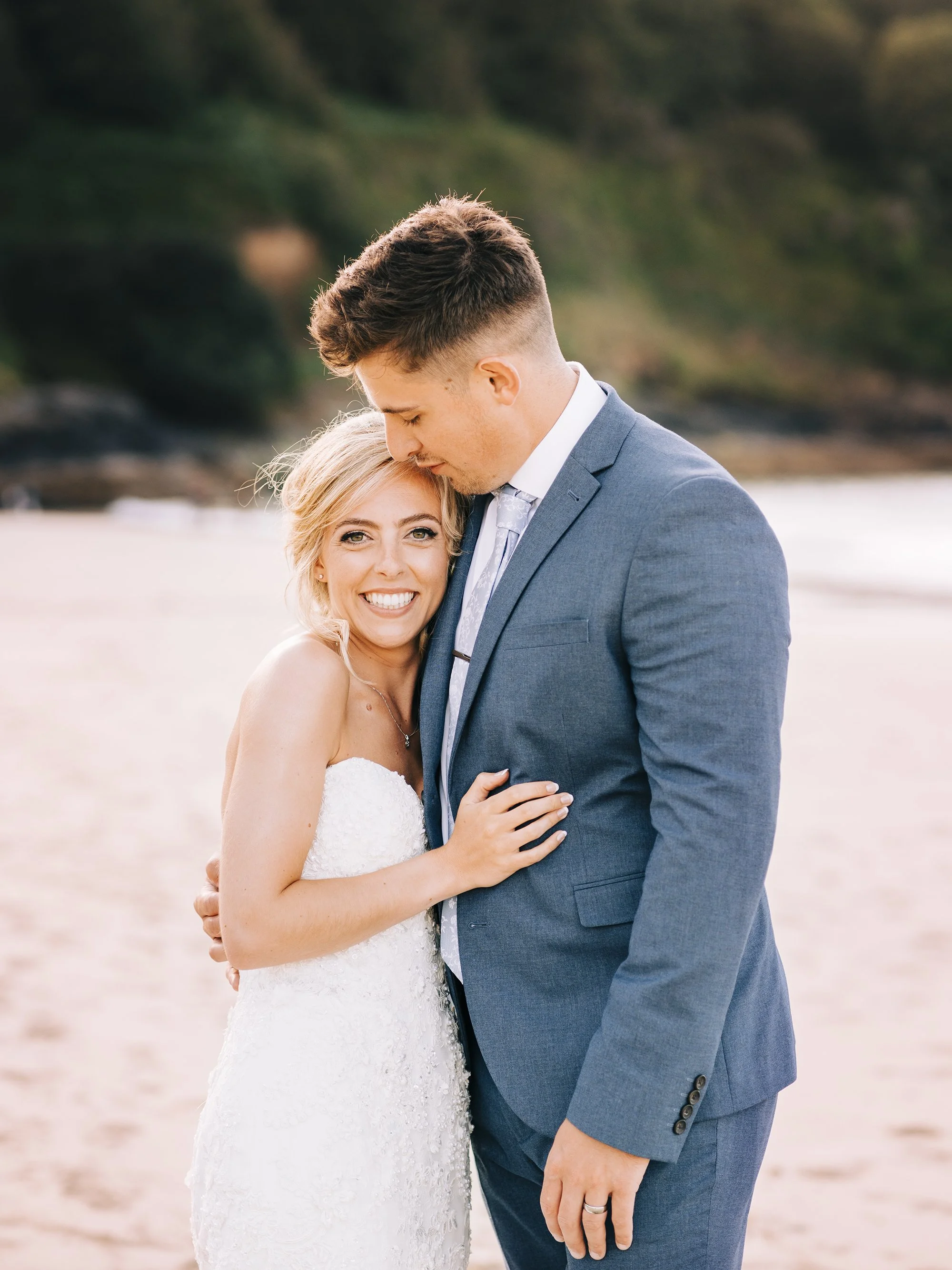 A happy bride and groom embrace on a beach during their wedding, with the bride smiling and the groom gently resting his forehead on her head.
