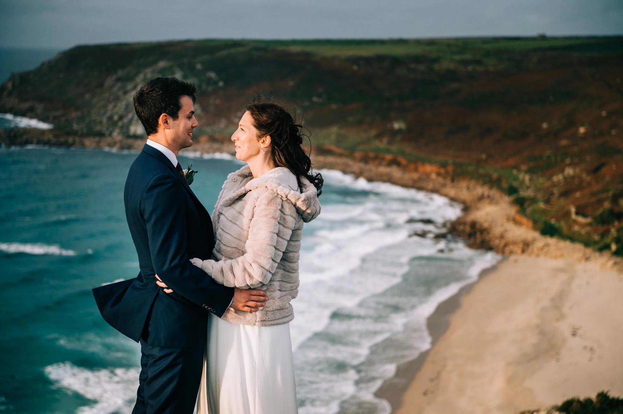 A newlywed couple stands on a beach, facing each other and smiling, with waves crashing on the shore and cliffs in the background.