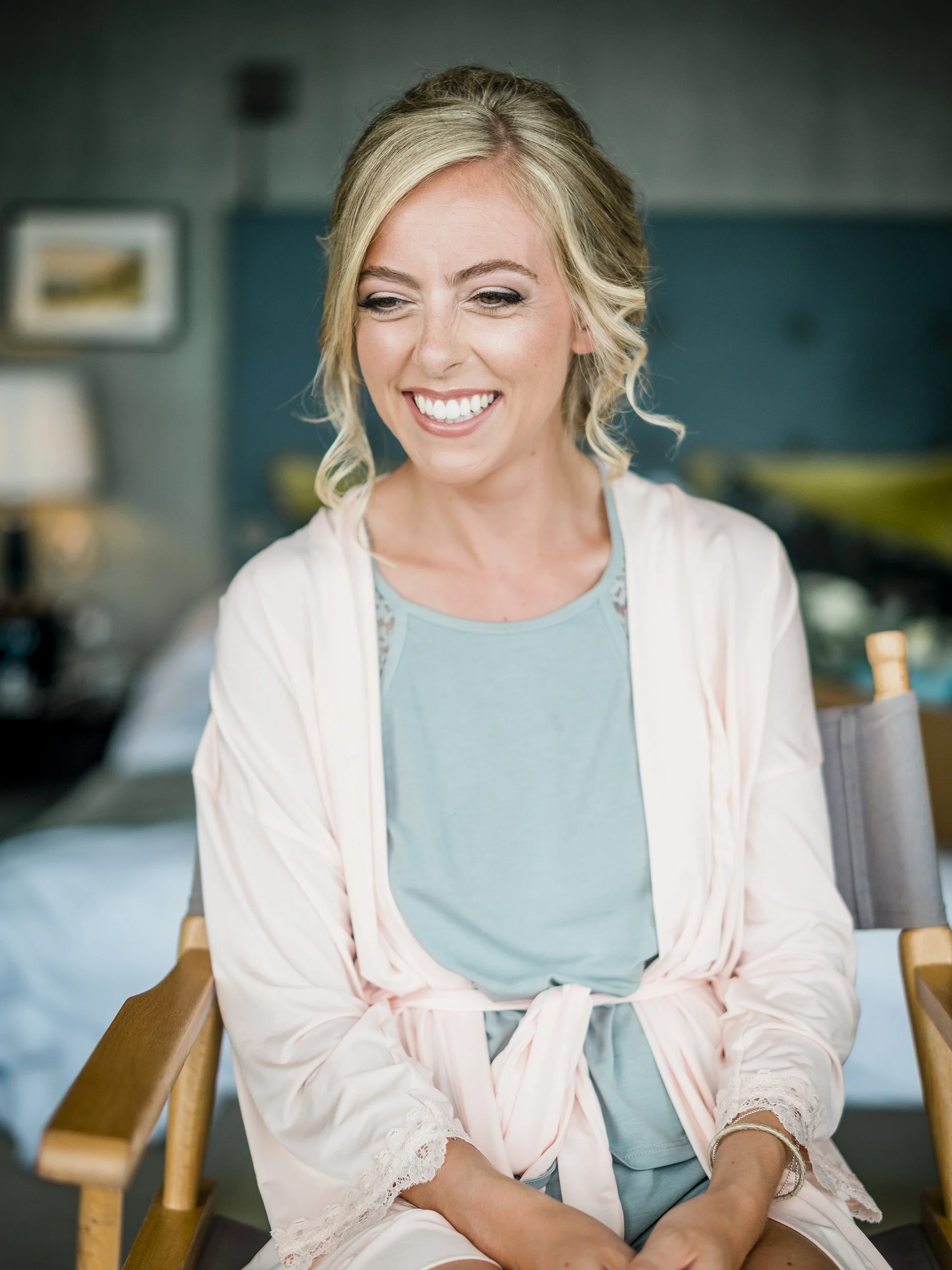 A woman with blonde hair styled in soft waves, smiling and looking to her left, wearing light blue pajamas with a matching robe tied at the waist, sitting in a wooden chair in a cozy room.