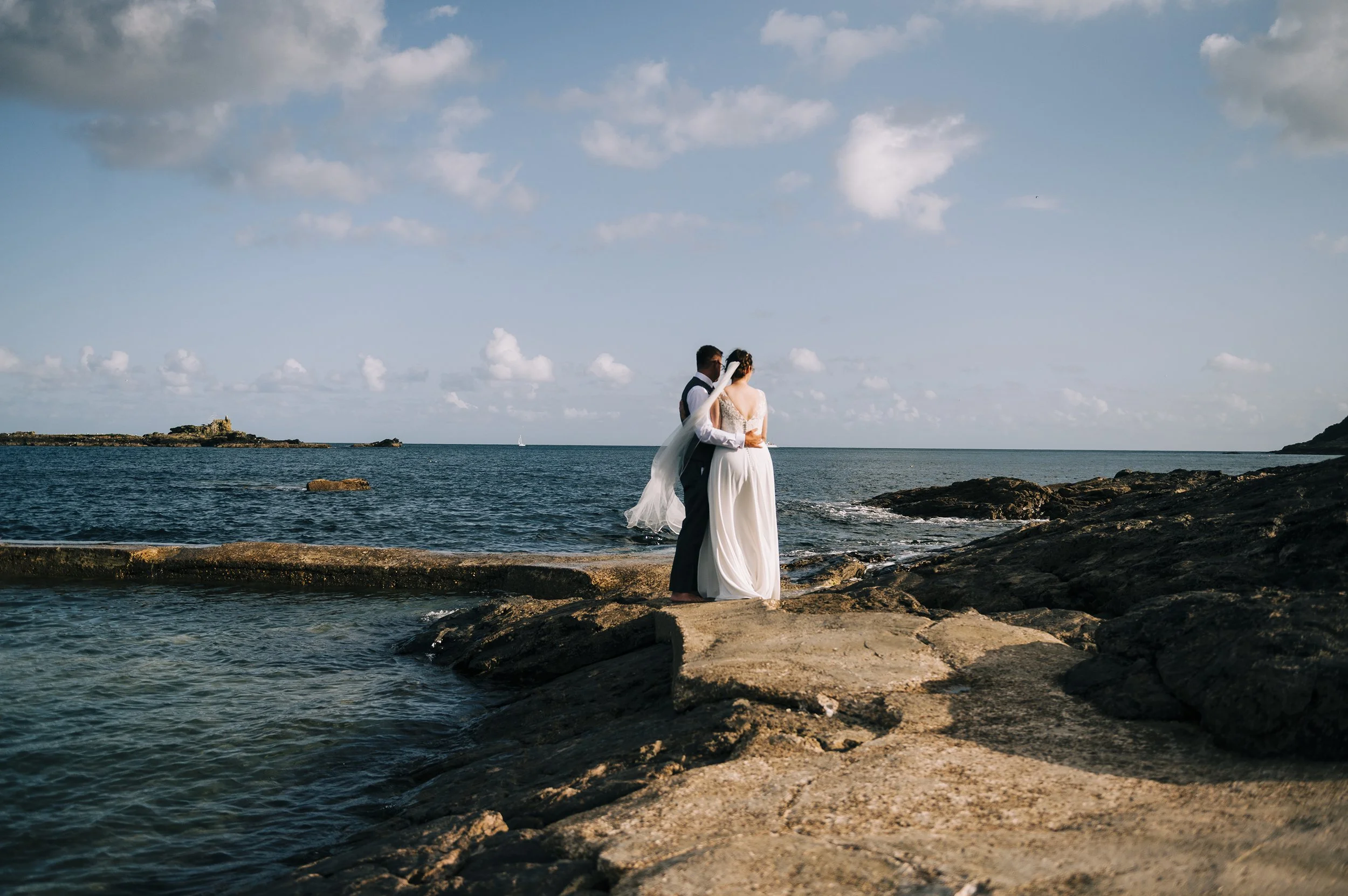 A bride and groom embrace on rocks by the ocean during sunset or late afternoon, with a cloudy sky and small island in the distance.