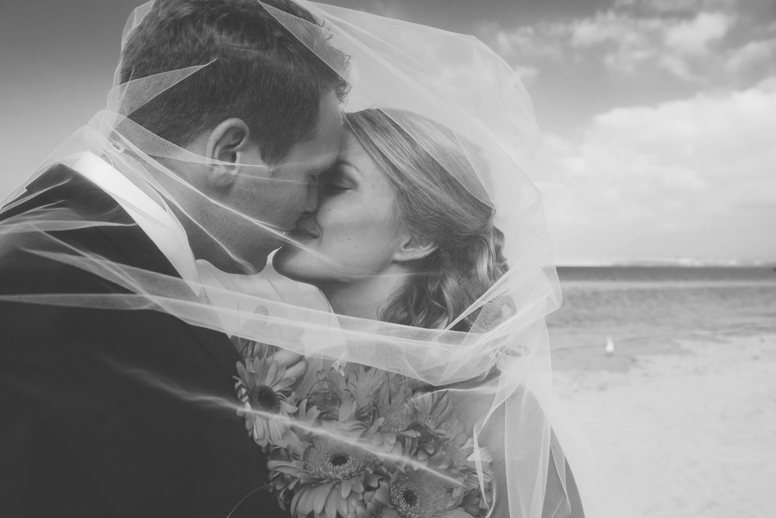 A black and white photo of a couple kissing on the beach, with the woman holding a bouquet of flowers and a sheer veil draped over both of them.