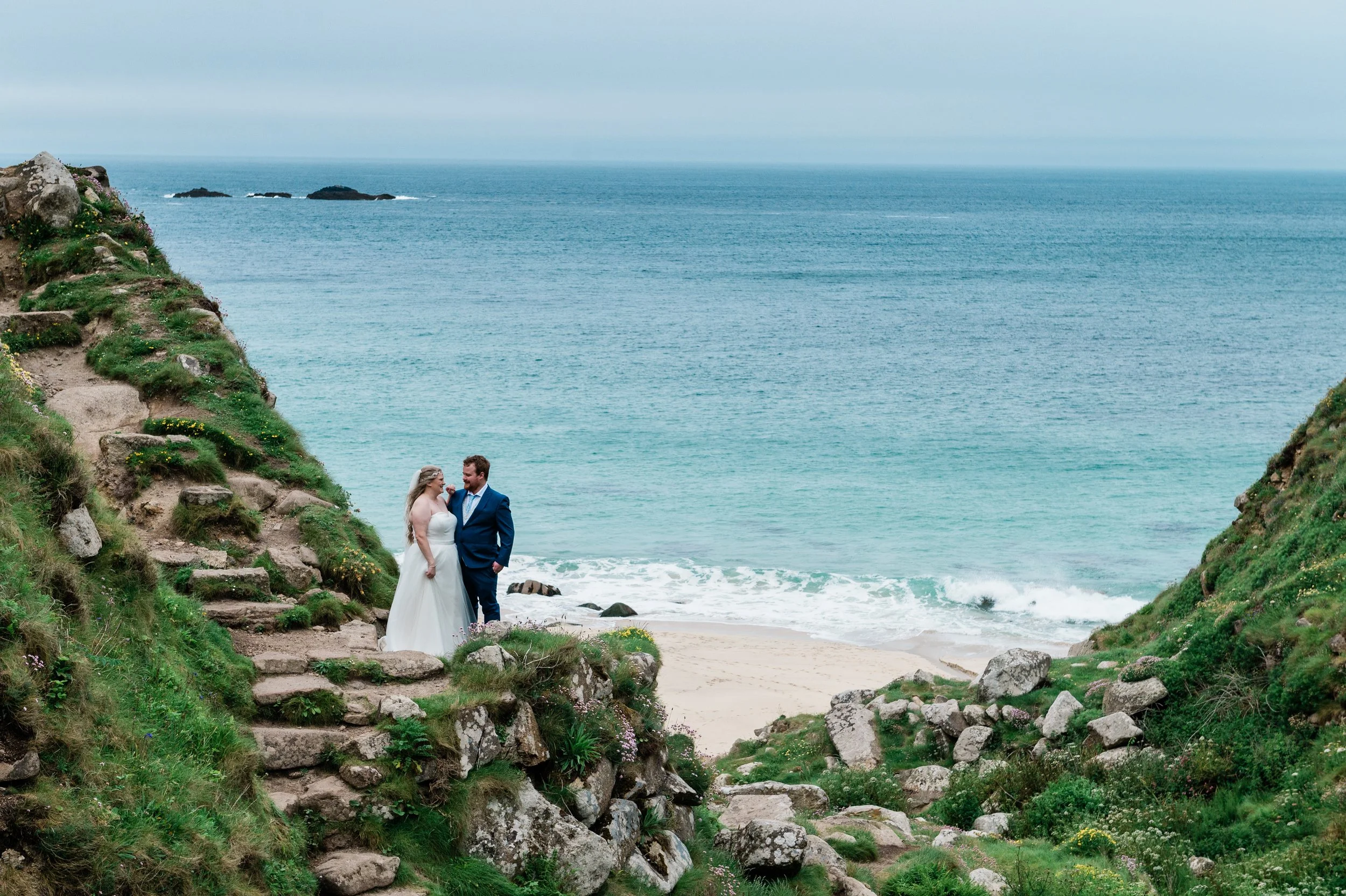A bride and groom standing on rocks near a beach with green grass and flowers, ocean waves in the background, with a cloudy sky.