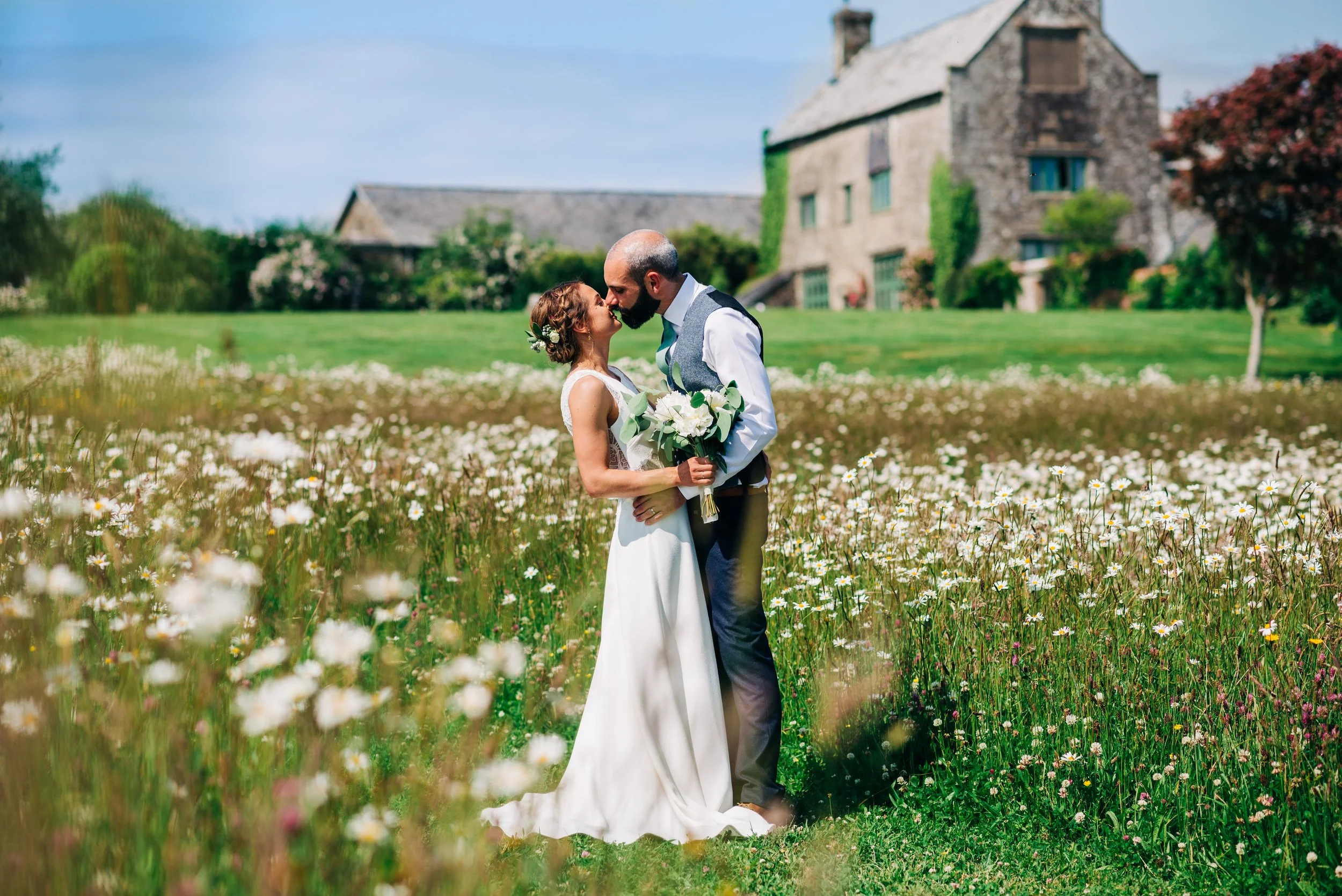 A bride and groom stand close together in a field of wildflowers, kissing. The bride holds a bouquet of white flowers. In the background is a large stone house with ivy and trees, under a blue sky with some clouds.