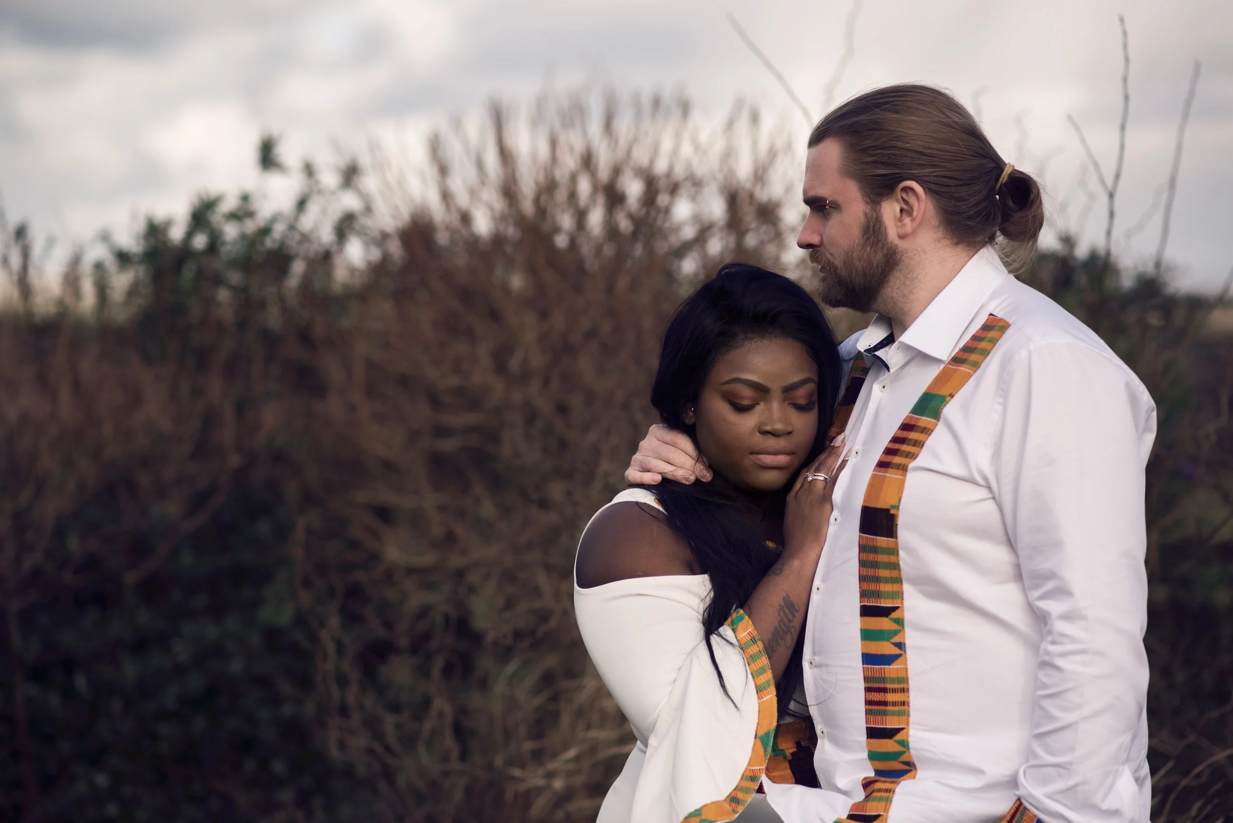 A couple embracing outdoors, the woman with her eyes closed and the man looking down at her, both dressed in white clothing with colorful patterns, standing in front of a background of trees and cloudy sky.