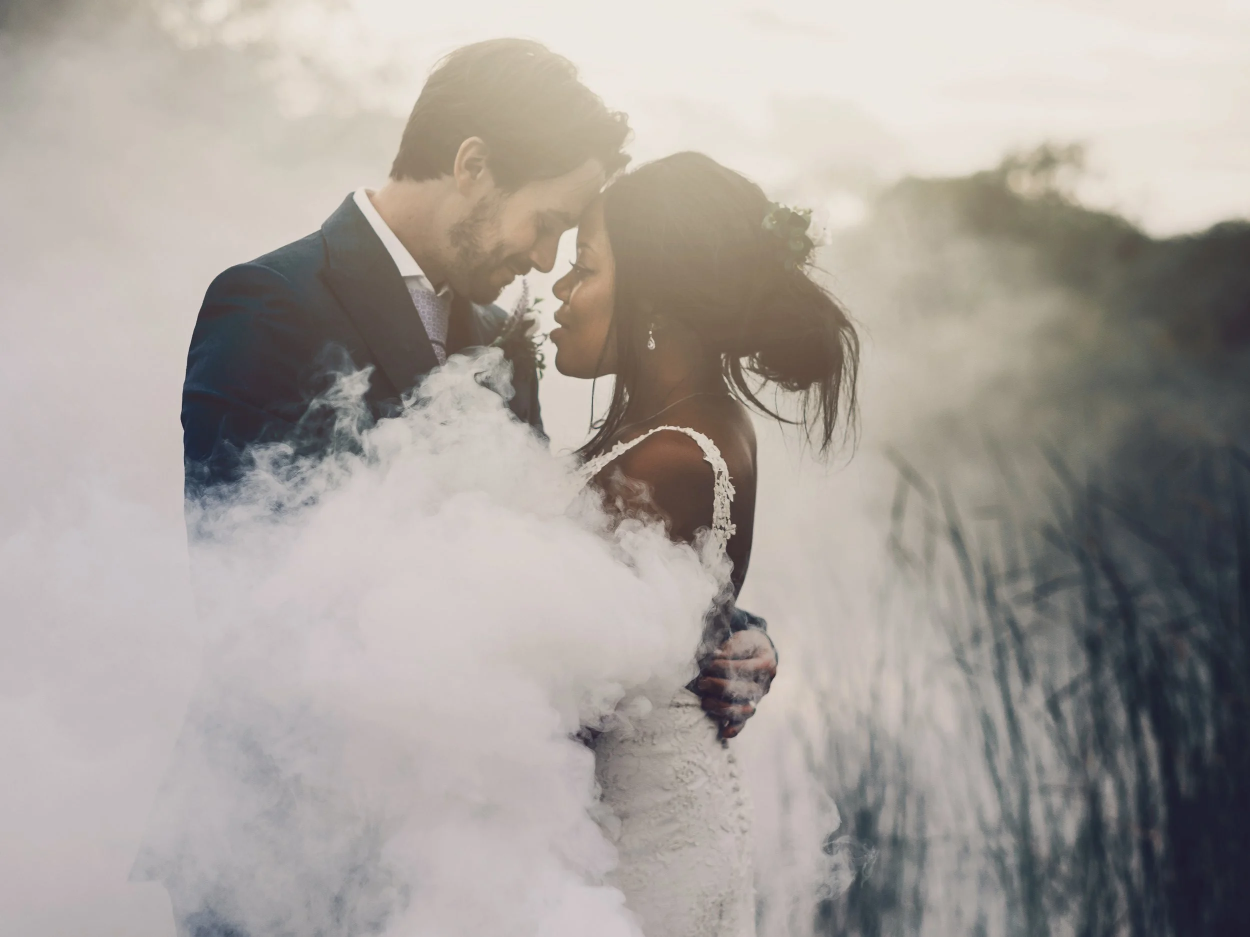A couple dressed in wedding attire, embracing with their foreheads touching, surrounded by smoke in an outdoor setting.