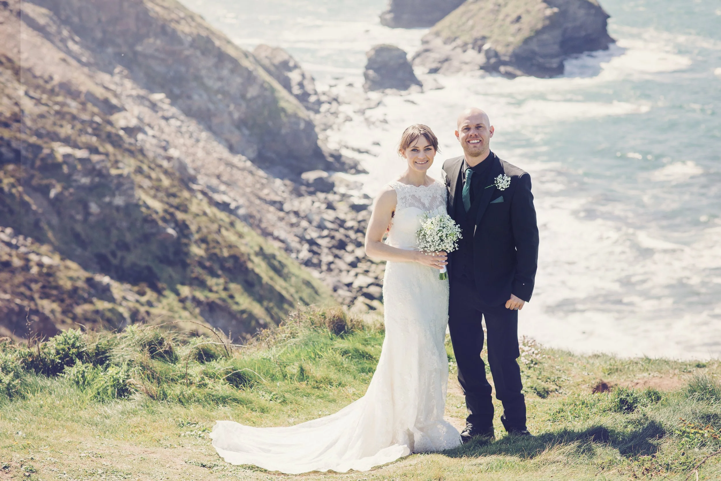 A bride and groom standing together outdoors by the coast, with cliffs and ocean in the background. The bride is wearing a white wedding gown and holding a bouquet of flowers. The groom is in a dark suit with a tie, smiling at the camera.