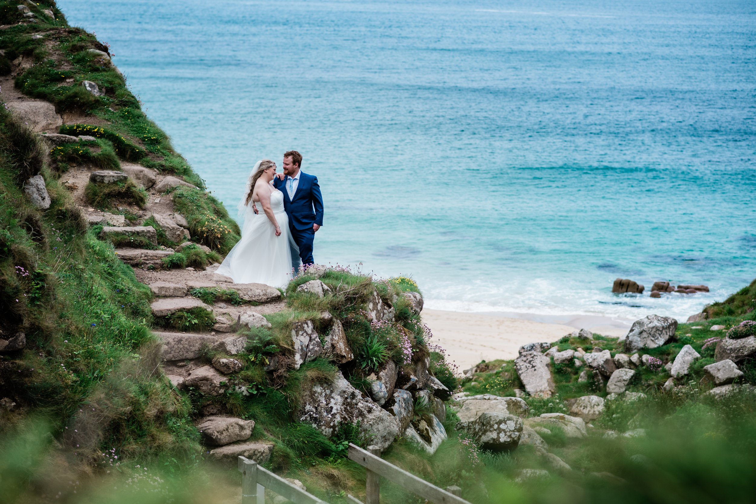 A bride and groom in wedding attire standing on rocky steps on a green hillside overlooking the ocean, with a sandy beach and waves in the background.