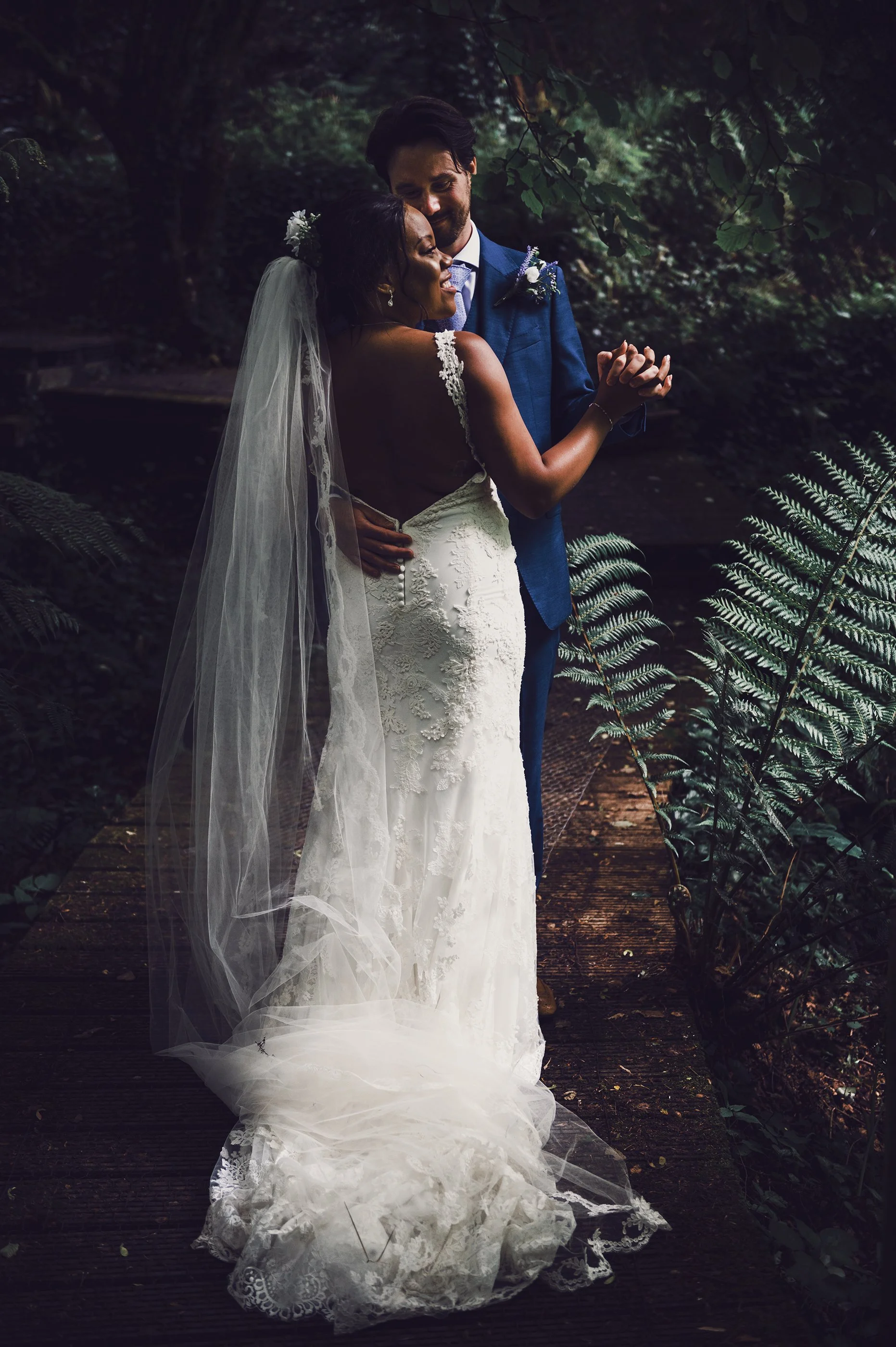 A bride and groom embrace on a wooden path in a forest, smiling and holding hands. The bride is wearing a white lace wedding dress and veil, and the groom is in a blue suit with a boutonniere. The scene is surrounded by lush green foliage.