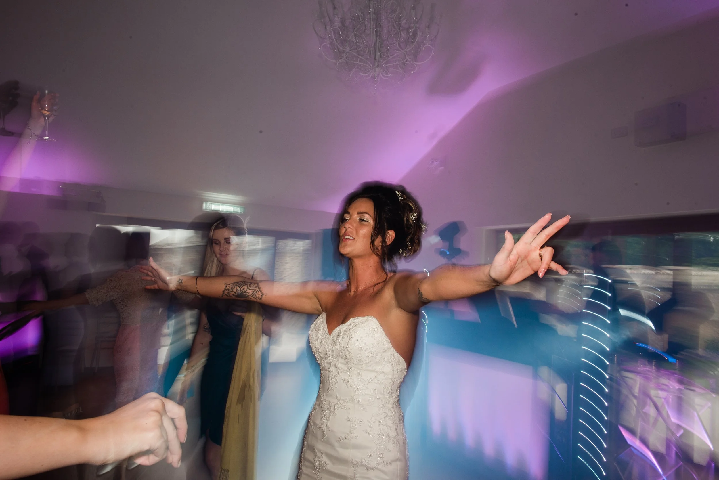 A bride in a white wedding dress with her arms outstretched celebrating at the wedding reception with purple and blue mood lighting.