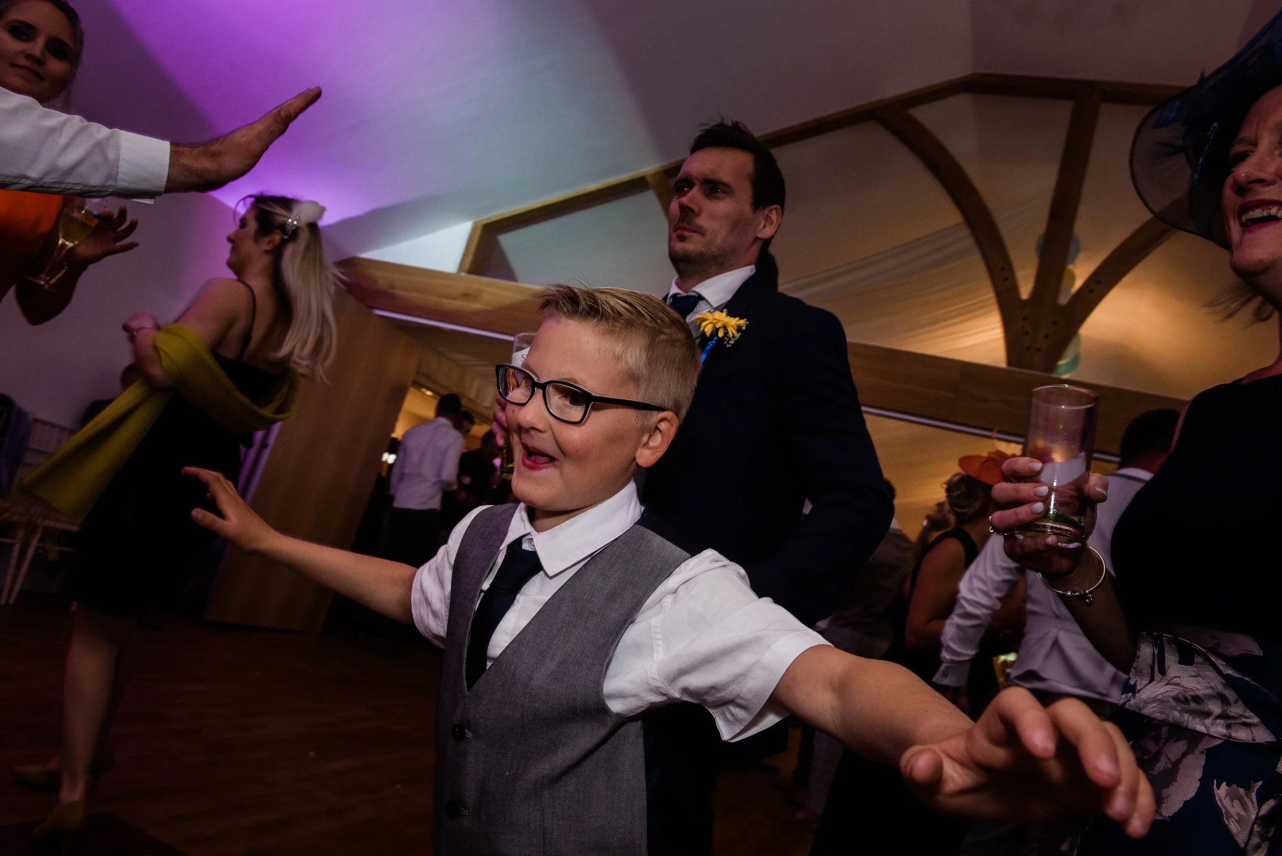 Children and adults dancing and celebrating at a party or wedding reception with purple lighting and a wooden ceiling beam in the background.