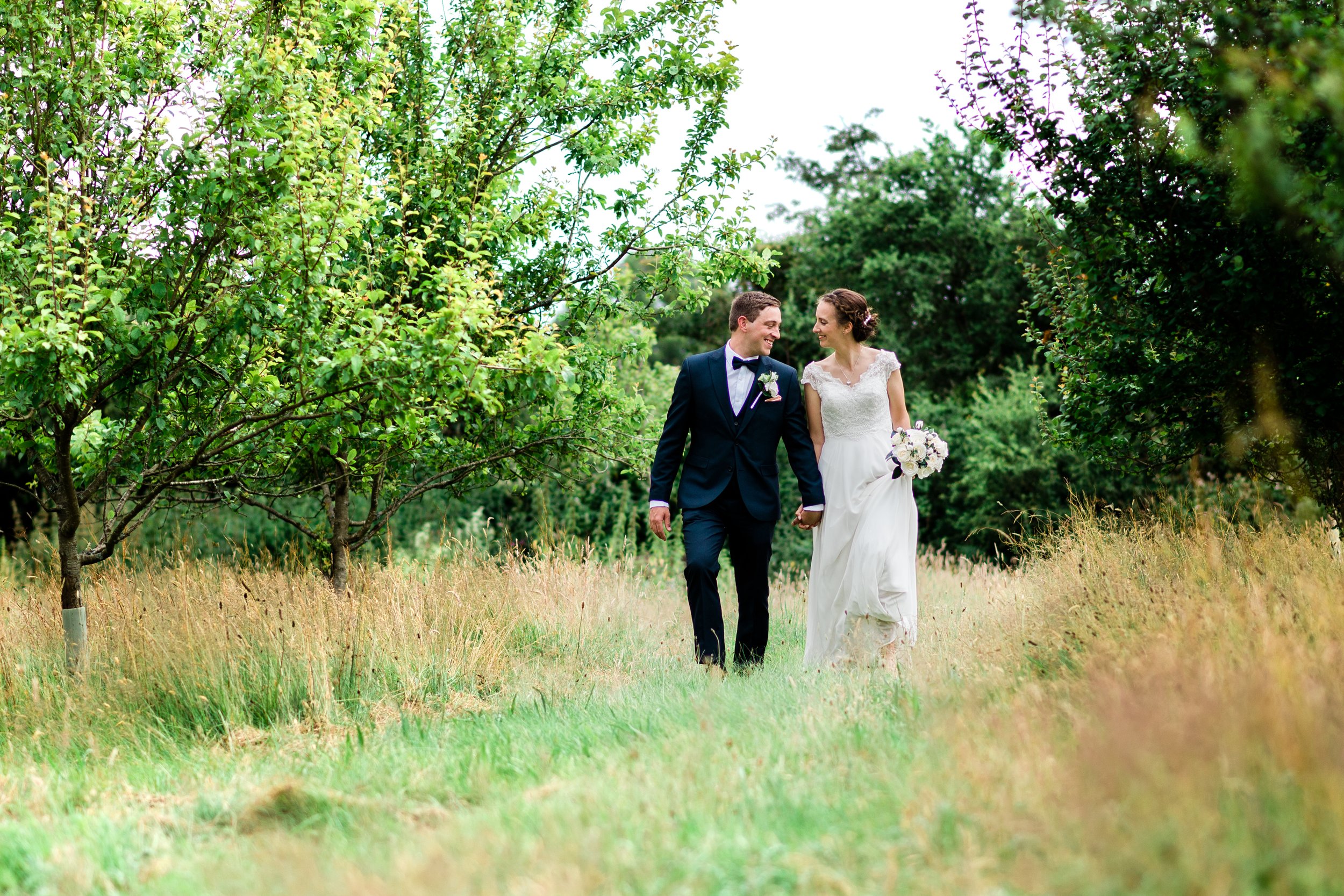 A newlywed couple walking hand in hand through a lush green outdoor setting, smiling at each other, with trees and grass around them.
