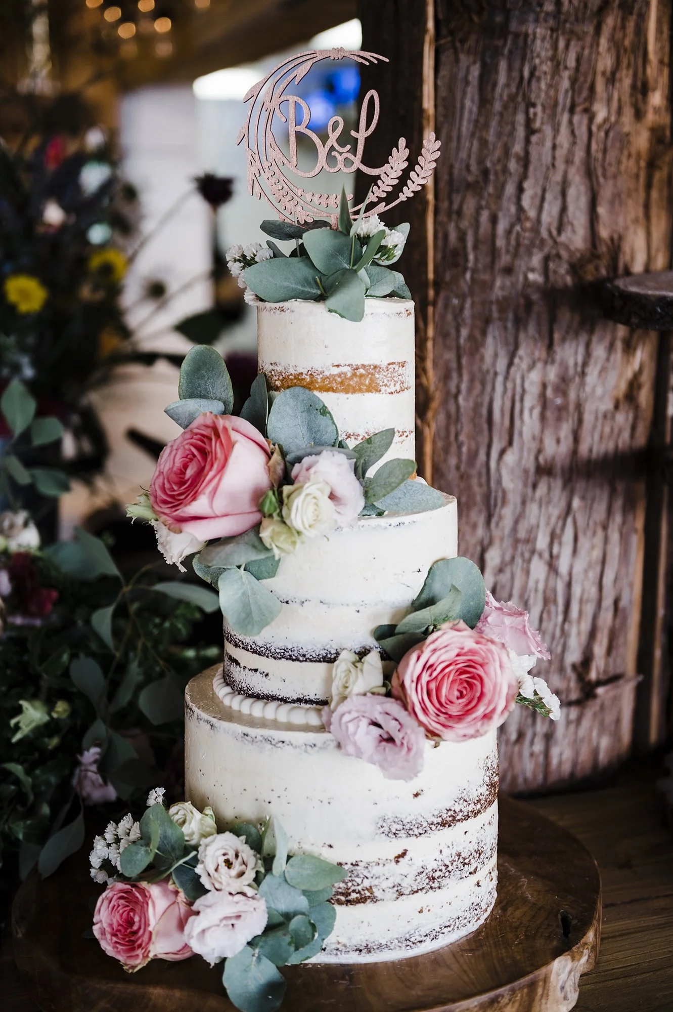A three-tier semi-naked wedding cake decorated with pink and white roses, eucalyptus leaves, and a pink topper with 'B&L' on it, placed on a wooden table against a rustic wooden wall.